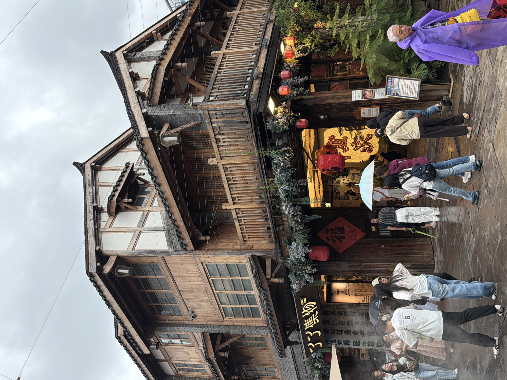In the Yuzhong District of China, a street scene unfolds under a cloudy sky, with traditional architecture dominating the frame. A multi-story wooden building, characteristic of local design, stands prominently. Its facade features multiple levels with balconies, wooden windows, and tiled roofs. Red lanterns and decorative floral arrangements adorn the building's exterior, suggesting a festive or cultural setting.
Several people are visible on the wet cobblestone street. Some are walking, others are conversing, and a few are holding umbrellas, indicating possible rain or misty conditions. One individual in the foreground is wearing a vibrant purple raincoat, adding a splash of color to the scene. The street appears to be a pedestrian area, with shops and businesses lining the building. The overall atmosphere is one of a bustling, yet somewhat subdued, urban environment, possibly during a day with inclement weather.