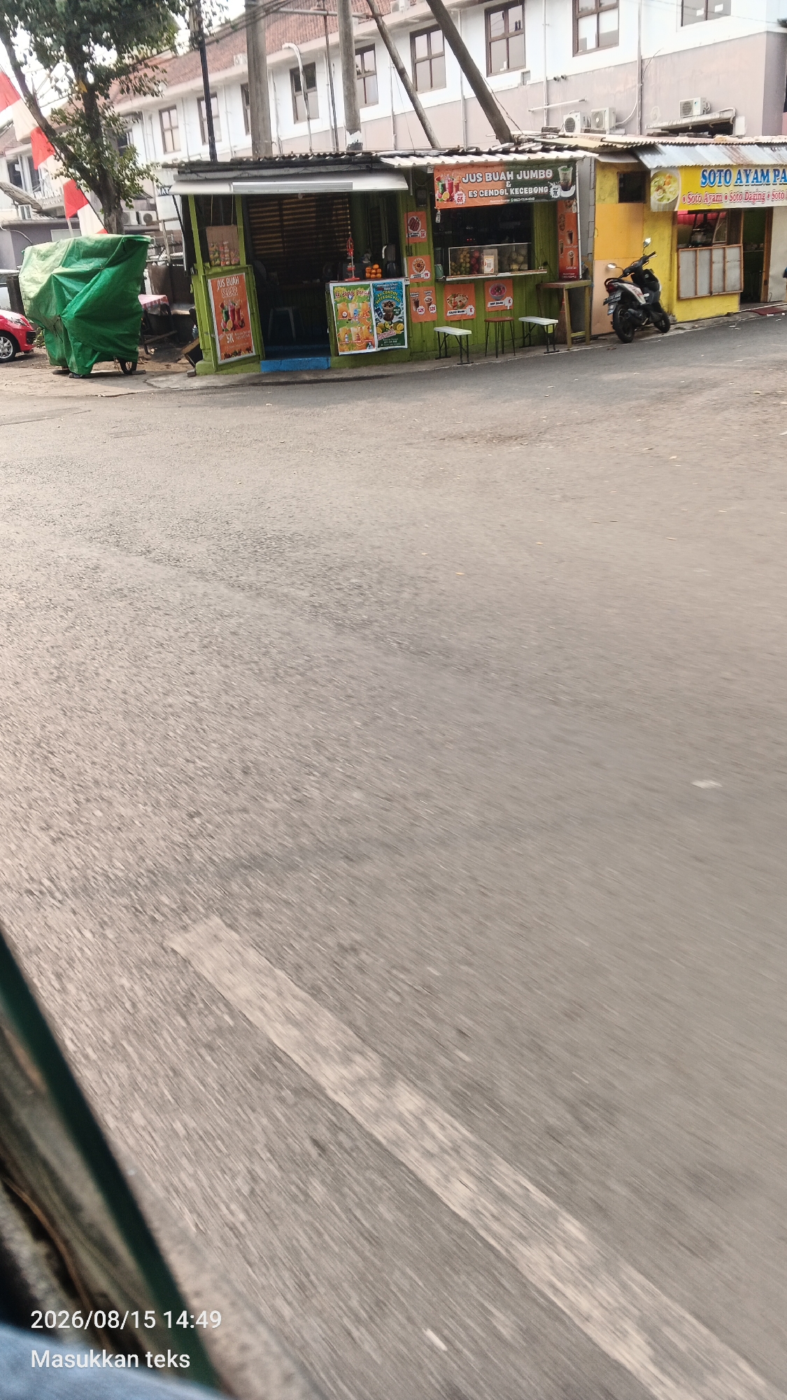 The image depicts a street scene in Malang, Indonesia, showing a row of small food stalls. The stalls appear to be made of corrugated metal with colorful signs. One stall, painted green, advertises "JUS BUAH JUMBO" (Jumbo Fruit Juice) and "ES CENDOL KECEBONG" (Kecebong's Cendol Ice). Another stall, painted yellow, has a sign for "SOTO AYAM PA" (Chicken Soto Pa). There are customers seated at small tables in front of the green stall. A motorcycle is parked in front of the yellow stall. The street in the foreground is paved and shows some wear. A green tarp covers an object on the left side of the image. The overall impression is a typical roadside food vendor area in a bustling Indonesian city.