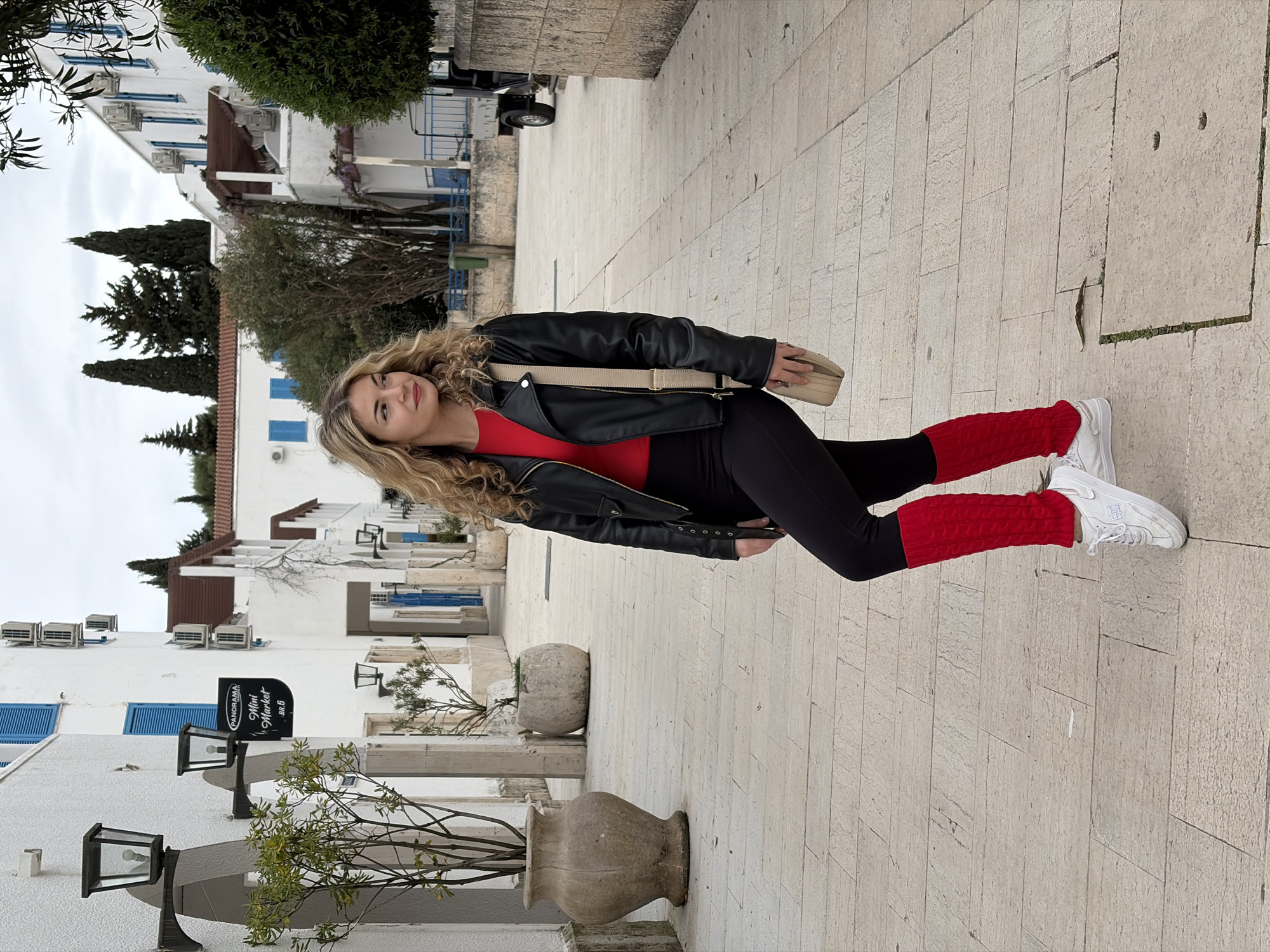 A young woman stands on a light beige paved street in Budva, Montenegro. She wears a black leather jacket over a red top, black leggings, red leg warmers, and white sneakers. A beige crossbody bag rests at her side. The background features white buildings with blue window shutters. Stone flower pots with plants line the street. A sign for "Panorama Mini Market B&G" is visible on one of the buildings. The sky is overcast.