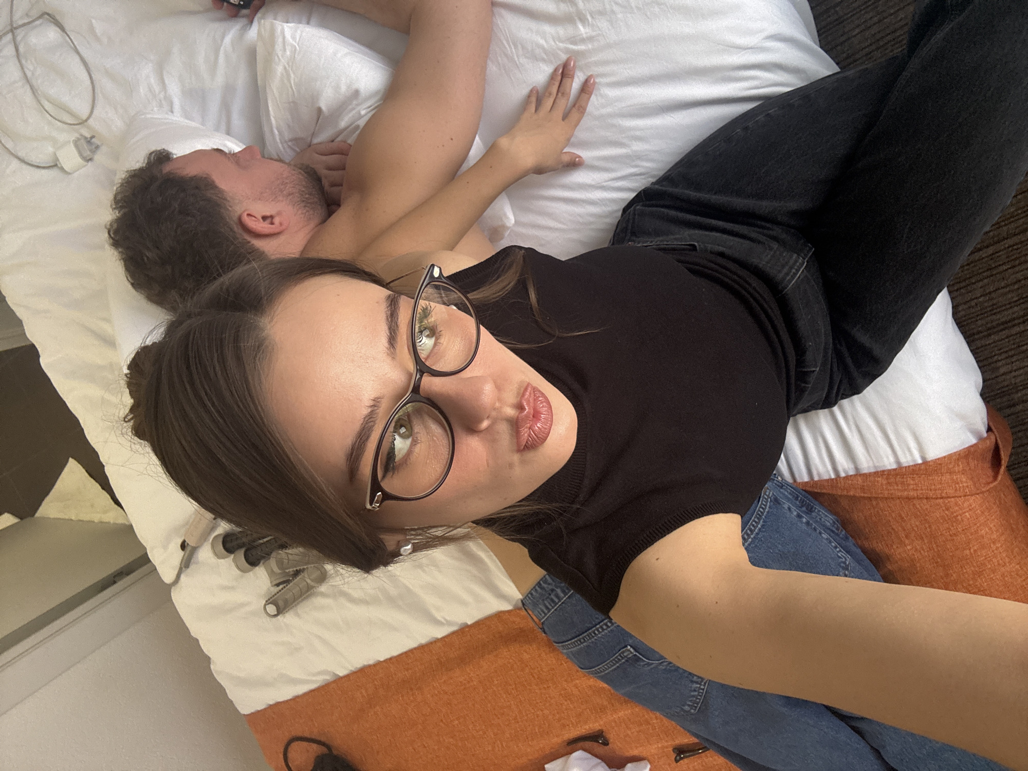 The image shows a woman in black jeans and a black top lying on a bed. She is wearing glasses and posing for a selfie. Behind her is a man lying on a pillow. The bedding is white with an orange accent. There are hair curlers and a phone charger on the bed near the pillows. The location is not specified.