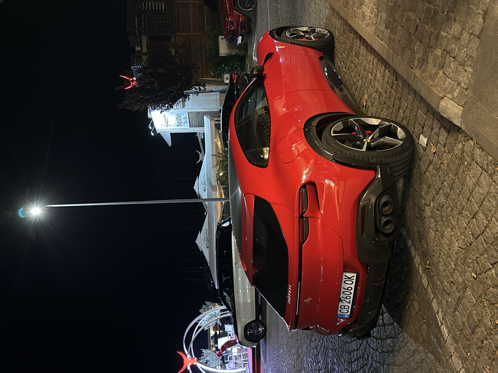 A bright red Ferrari Purosangue is parked on a cobblestone street at night, facing away from the camera, with its rear lights and license plate illuminated. The car's license plate reads "CB 2606 OK" and it is a Bulgarian plate. To the left of the Ferrari, a white SUV with a black roof is parked. Further in the background, there are tents and buildings, one of which has a sign that reads "THEATRO". A street lamp is overhead, casting a bright light. The scene appears to be in Sozopol, Bulgaria, a coastal town known for its historic architecture and picturesque harbor.