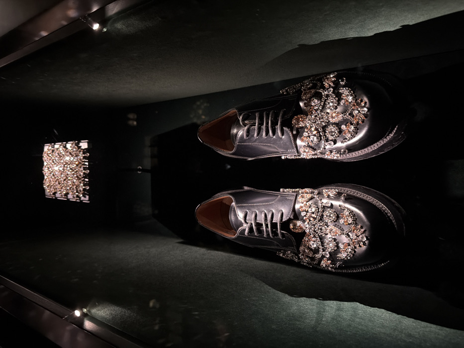 A pair of black leather derby shoes, heavily embellished with intricate patterns of clear and amber-toned crystals across the toe box and vamp, are centrally displayed within a dark, recessed exhibit. The shoes feature polished leather uppers, dark laces, and thick, prominent soles. The display environment consists of dark, possibly fabric-lined walls, exhibiting a deep green or teal hue under focused illumination from discreet spotlights. A rectangular, multi-tiered chandelier composed of numerous small, light-colored crystals is suspended from the dark ceiling, casting light and reflections. No individuals are present. This scene depicts a luxury fashion presentation, consistent with a high-end boutique like Balmain, located in Paris, France, likely at a flagship store such as the one on Rue Saint-Honoré.