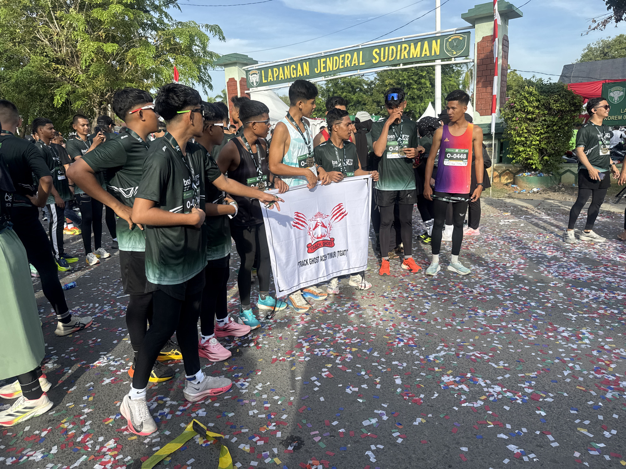 This is a photograph taken at the Lapangan Jenderal Sudirman sports field in Lhokseumawe, Indonesia. A group of young men, presumably runners from a team named "TRACK GHOST ACEH TIMUR (TGAT)," are gathered in front of an entrance archway. They are wearing matching dark green athletic shirts with some individuals sporting colorful singlets. Many have medals around their necks, and some are holding a white banner with red graphics. The ground is covered with confetti, suggesting a recent event or celebration. The men are of various ethnicities and appear to be of diverse ages, all casually interacting or posing for the camera. The background features a large tree, a brick and concrete archway with Indonesian flag, and a blue sky.
