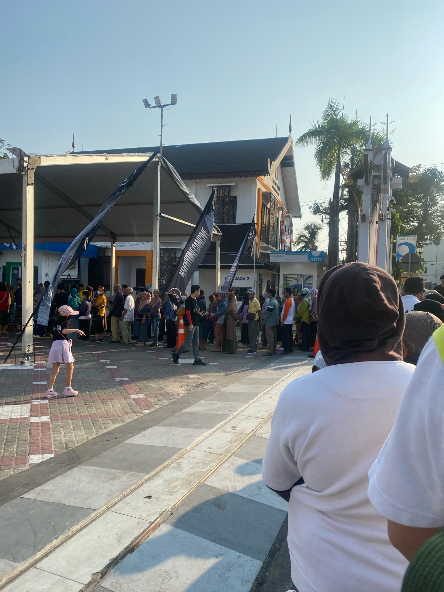 The image depicts an outdoor scene in Banjarmasin, Indonesia, featuring a crowd of people lined up outdoors under a clear, bright sky, suggesting it is daytime. A building with white and orange accents is visible in the background, with signs indicating "Bank Kalsel" and "ATM." Palm trees are also present in the background, indicative of a tropical or subtropical climate. Several black banners with white text are suspended from poles, some of which have the hashtag "#WEVERGONCAGIVE" visible.
In the foreground, a young girl wearing a pink cap and a light purple skirt is standing on a patterned walkway, appearing to be interacting with someone off-camera or performing a dance. Behind her, a line of people, many of whom appear to be wearing casual attire and headscarves, are queuing. The ground is paved with a combination of brick and concrete. The lighting suggests a sunny day, with distinct shadows cast on the ground. The overall impression is of an organized gathering or event.