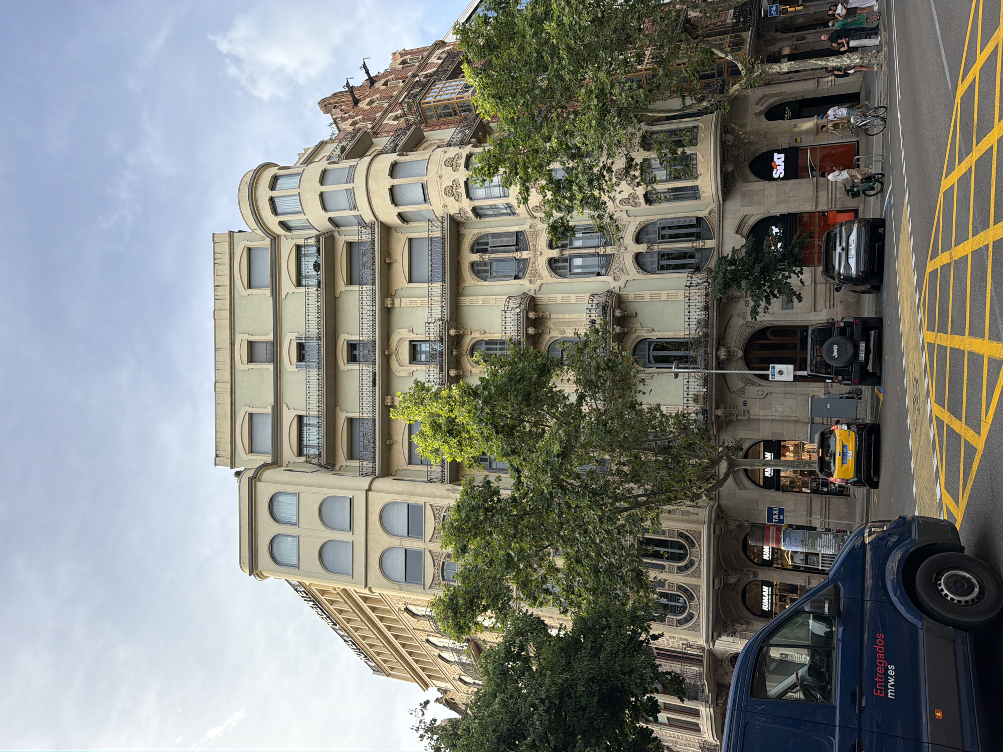 This is an eye-level, medium shot captured on a slightly overcast day in Barcelona, Spain. The image is dominated by the facade of a large, multi-story building with a light green and beige color scheme, featuring ornate architectural details such as balconies with wrought-iron railings and arched windows. To the left, a portion of an adjacent building with a lighter beige facade and arched windows is visible. Trees with lush green foliage line the street in front of the buildings.
In the foreground, a dark blue delivery van with "Entregados mrw.es" written in red on its side is parked on the left. Several cars are also present: a yellow taxi, a black Jeep, and a dark grey SUV are stopped at a street intersection marked with yellow crosswalk lines. Several people are standing on the sidewalk near the buildings, some appearing to be pedestrians and others interacting near a Sixt rental car office, identified by its orange signage. Two bicycles are also visible on the sidewalk. The sky above is a light blue with scattered white clouds. The scene depicts a typical urban streetscape in Barcelona.