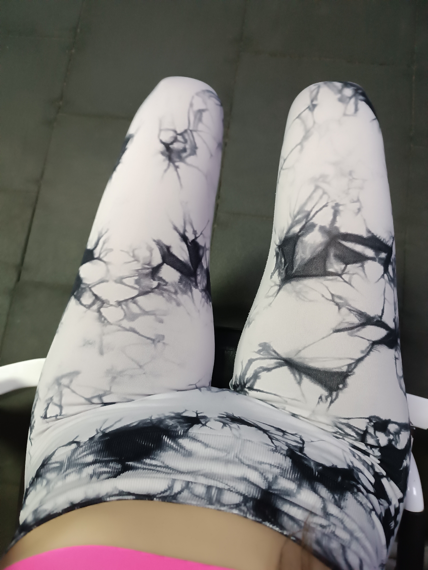The image shows a person's legs wearing white leggings with a black tie-dye pattern. The person is sitting on a white chair. The background is a blurry gray surface, possibly a floor or wall. The location is Unknown, Unknown. A pink garment is partially visible at the bottom of the image.