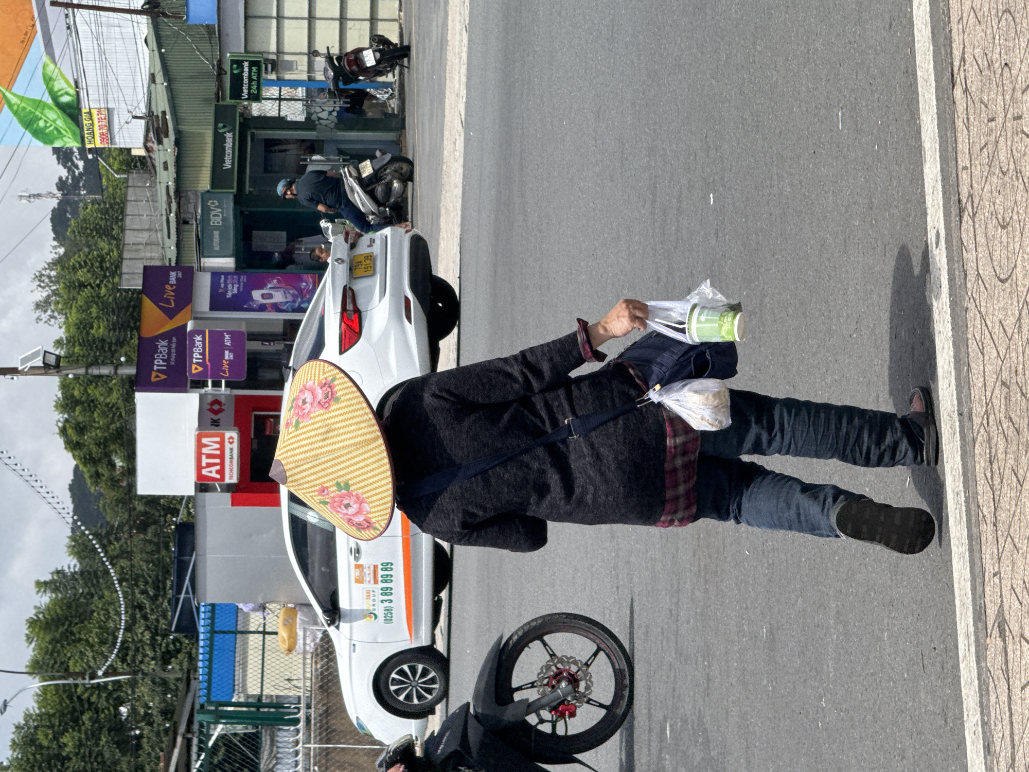 A person wearing a conical hat adorned with pink flowers and dark clothing walks across a street in Da Lat, Vietnam. The person carries a plastic bag containing a green cup and another bag. A white car with the number plate "79A 191.39" is visible in the background, along with several motorbikes. Buildings with signs for banks, including TPBank, BIDV, and Vietcombank, are also present. The street is paved with asphalt and bordered by a sidewalk with a tiled pattern. Trees and a hilly landscape are visible in the distance. The sky is partly cloudy.