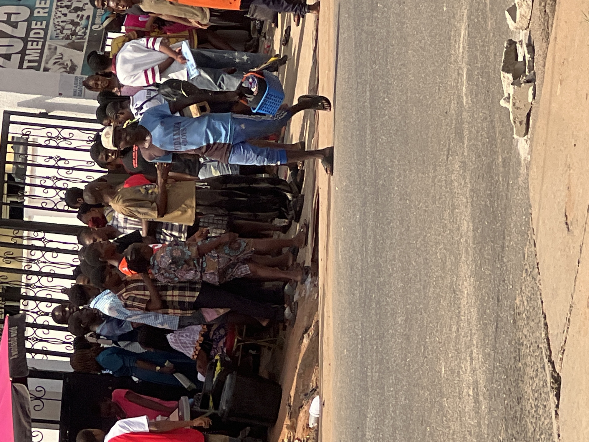 The image depicts a street scene in Nkpor, Nigeria, featuring a dense crowd of people gathered near a building with a poster on its exterior. Many people are standing in close proximity to each other, with some holding objects. The environment includes a paved road adjacent to the crowd, and the overall impression is that of a busy public space. A fence is visible between the crowd and the building.