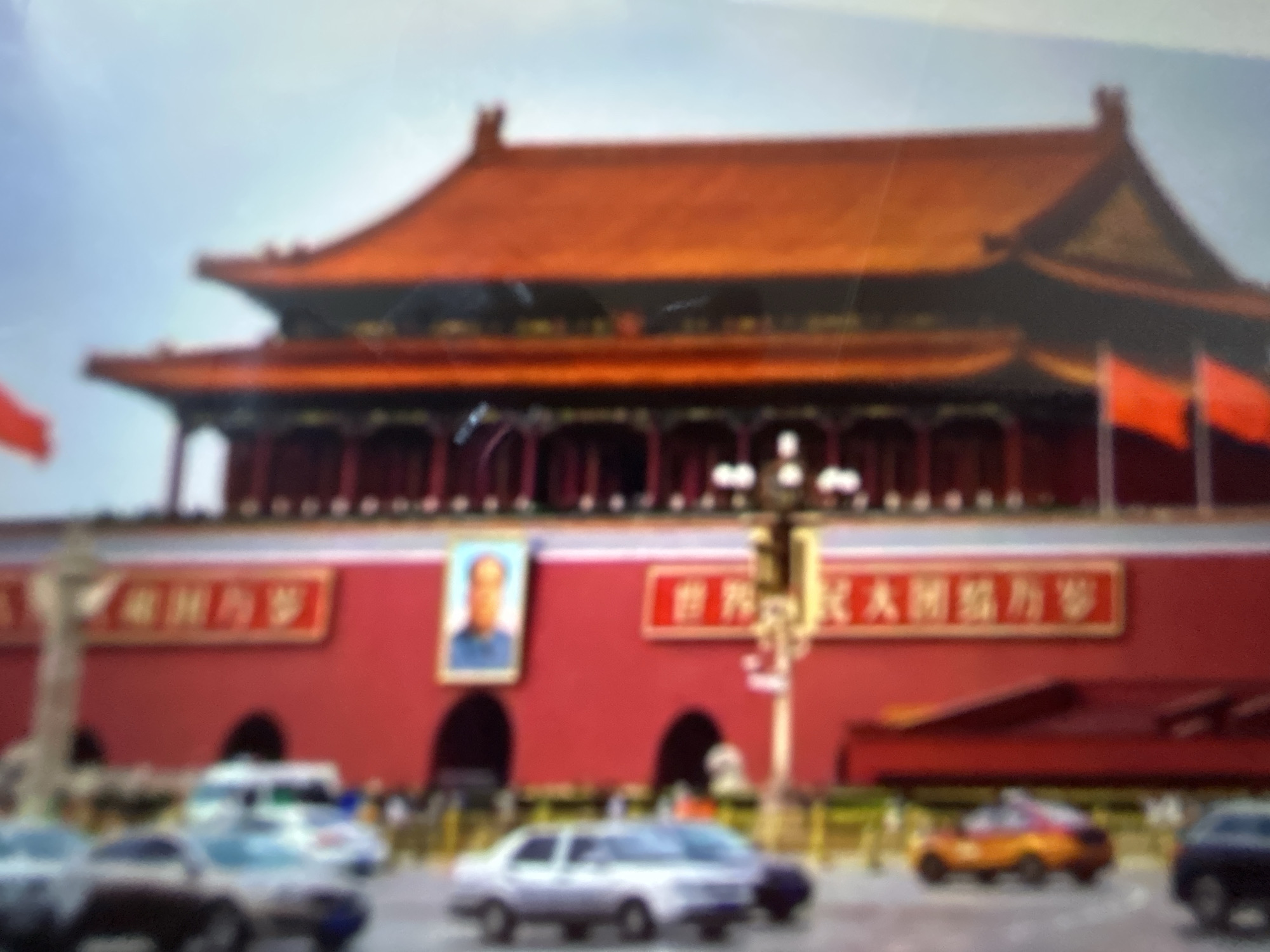 The provided image is a vertical, out-of-focus photograph of a large red building with ornate orange and brown roofing, characteristic of traditional Chinese architecture. The building features multiple levels and decorative elements. In front of the building, there is a street with several vehicles, including a white van and a yellow car. There are also red banners with Chinese characters displayed on the building's facade. A portrait of a person is visible on the wall. The sky is light blue. The image does not depict Gelendzhik, Russia; the architectural style and signage suggest a location in China, likely Beijing given the building's resemblance to the Tiananmen Gate.