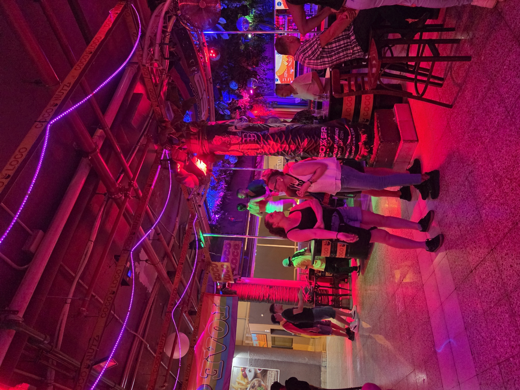 The image depicts the interior of a bar or nightclub in San Bartolomé de Tirajana, Spain, illuminated by vibrant pink and purple neon lights. The scene is bustling with people standing and socializing. In the foreground, two women are conversing near a bar area adorned with decorative elements, including what appears to be a faux palm tree wrapped in black material with printed text. To the left, a group of men are standing near a food advertisement banner. Further into the background, more patrons are visible, some standing at the bar and others mingling. The environment features a textured floor, ceiling structures with exposed pipes and lighting, and various decorative items such as disco balls. The overall atmosphere suggests a lively evening entertainment venue.
