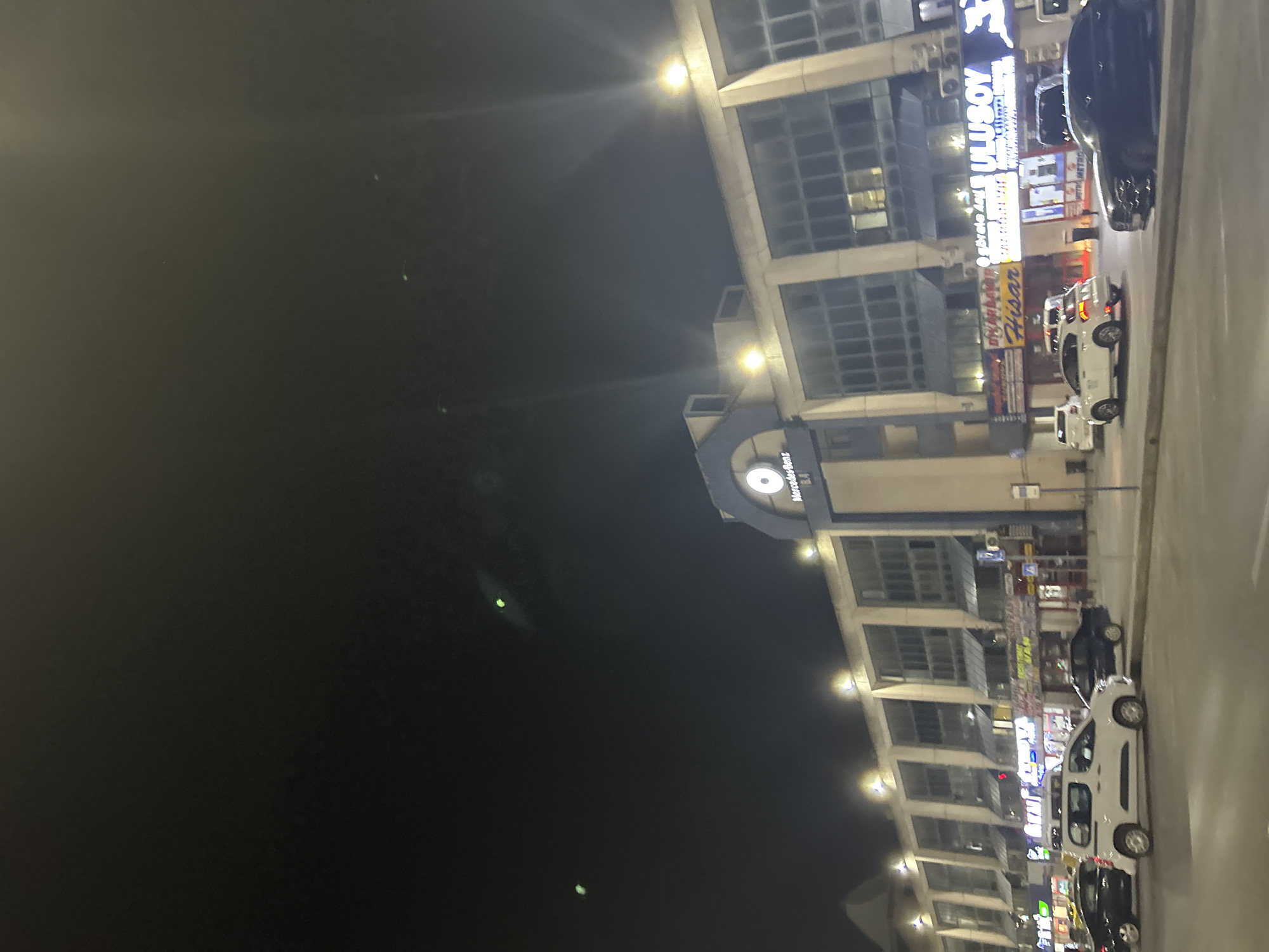 This media file captures a nighttime urban scene in Bayrampaşa, Turkey, viewed from a slightly elevated perspective. The image is oriented sideways, but if rotated 90 degrees counter-clockwise, it shows a multi-story commercial building on the right, brightly lit, with a street running along its base.
The dominant subject is a modern-looking building featuring numerous large glass windows and concrete or light-colored facade elements. Its exterior is illuminated by several bright spotlights, creating distinct lens flares in the dark sky. Along the street directly in front of the building, several vehicles are parked; these include a number of white vans or cars and at least one dark-colored car.
The upper left portion of the image is a dark night sky, with a few faint, distant lights or reflections visible. The atmosphere is calm, indicative of a typical urban evening.
Visible text on the illuminated signs along the building's facade provides location cues. Prominently displayed is "ULUSOY" in large white letters on a blue background. Below this, smaller text reads "MERCEDES METRO". Another sign features the word "HİSAR". Further down the building, a blue sign with a circular white logo and the text "Mercedes-Benz" is discernible. These details, combined with the urban architecture, confirm the commercial setting in Bayrampaşa, Turkey. No people are clearly visible, and the activity is limited to the presence of parked vehicles and illuminated businesses.