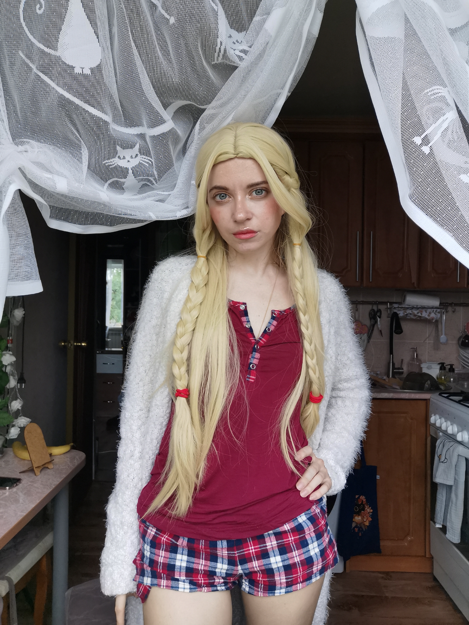A young woman with blonde braided hair and makeup poses in a dimly lit room. She is wearing a red shirt and plaid shorts, with a white fuzzy cardigan. The background shows a kitchen with cabinets, a stove, and a window. The room appears to be a domestic setting, possibly an apartment or house, and there is no indication of it being located in a specific city or region.