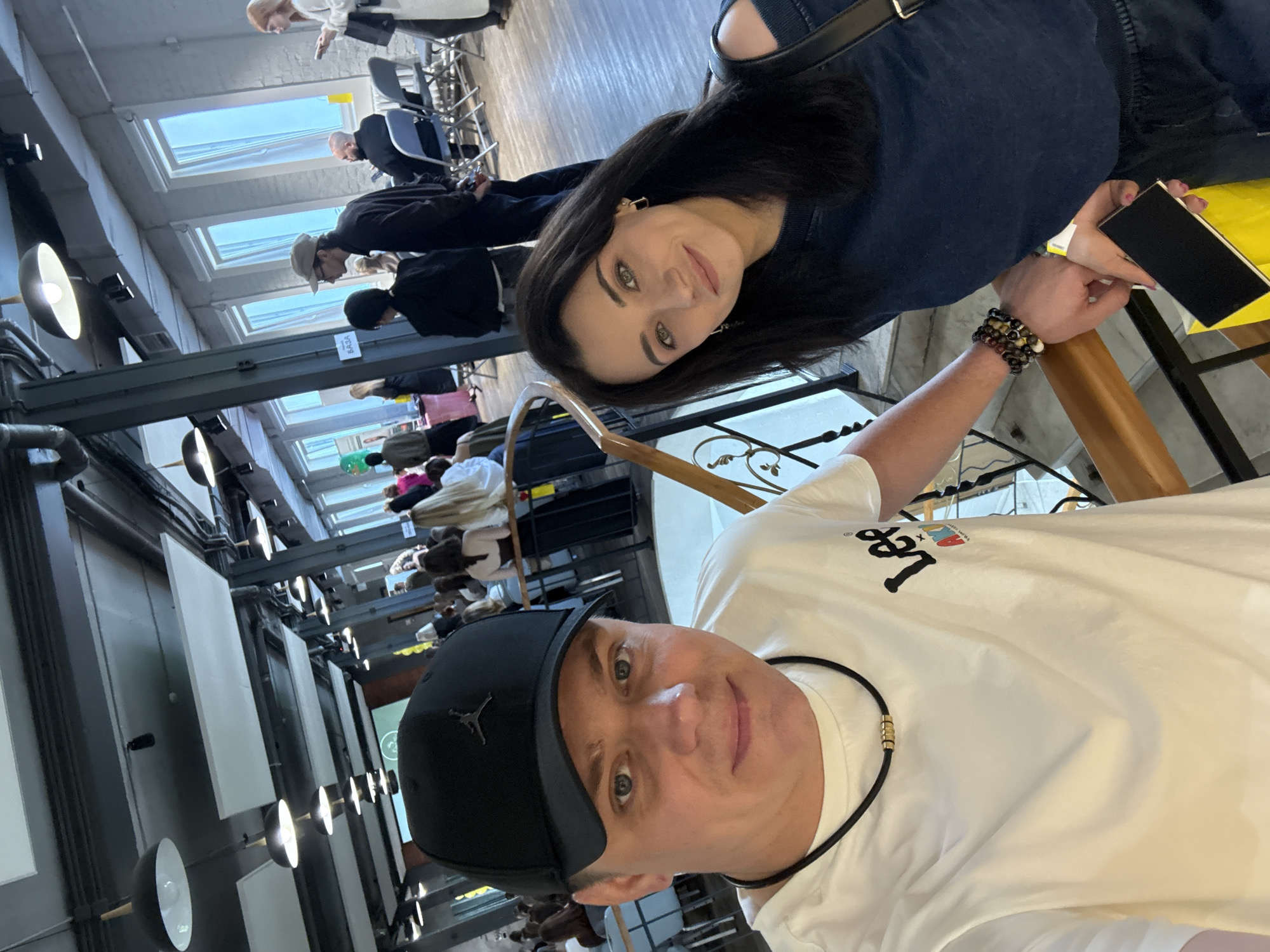 The image shows a man and a woman taking a selfie from an elevated position within a large, brightly lit indoor space. The man, positioned on the left in the foreground, wears a black baseball cap with a Jumpman logo, a white t-shirt with a "Lee" logo, and a black necklace with gold cylindrical beads. The woman, on the right, has dark hair and wears a dark blue off-the-shoulder top, multiple beaded bracelets on her right wrist, and holds a black phone or wallet and a yellow bag. Both are looking towards the camera with slight smiles.
They are leaning against a wooden handrail with black metal balustrade, overlooking a spacious event area below. The background reveals an interior with an industrial aesthetic, featuring exposed dark metal beams and columns, numerous black pendant light fixtures, and white rectangular acoustic panels on the ceiling. A prominent wall in the distance is lined with multiple tall, vertical windows, providing natural light. Below, a crowd of people is visible, some standing and interacting, others seated on gray folding chairs at tables. A sign partially visible in the background displays the Cyrillic word "БАЗА" (BAZA), and a projector screen shows a green circular logo. The architectural characteristics and the "BAZA" sign indicate the location is the BAZA creative cluster and event space, situated at Fontannaya 2a in Vladivostok, Russia, likely during a public event or gathering.