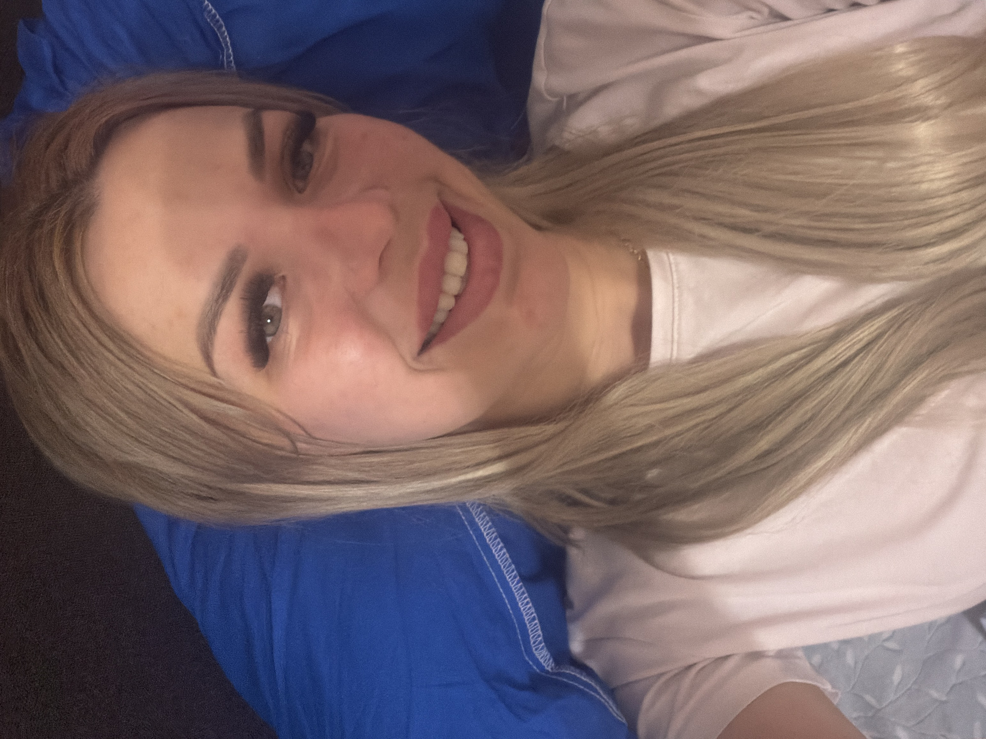 A blonde woman with light skin and long blonde hair is lying down, looking at the camera. She is wearing a light pink t-shirt. Her makeup includes false eyelashes and lip gloss. She is on a bed with blue and white patterned sheets and a blue blanket. The image was taken indoors, likely in a bedroom. There is no discernible indication of the location being Kyiv, Ukraine.