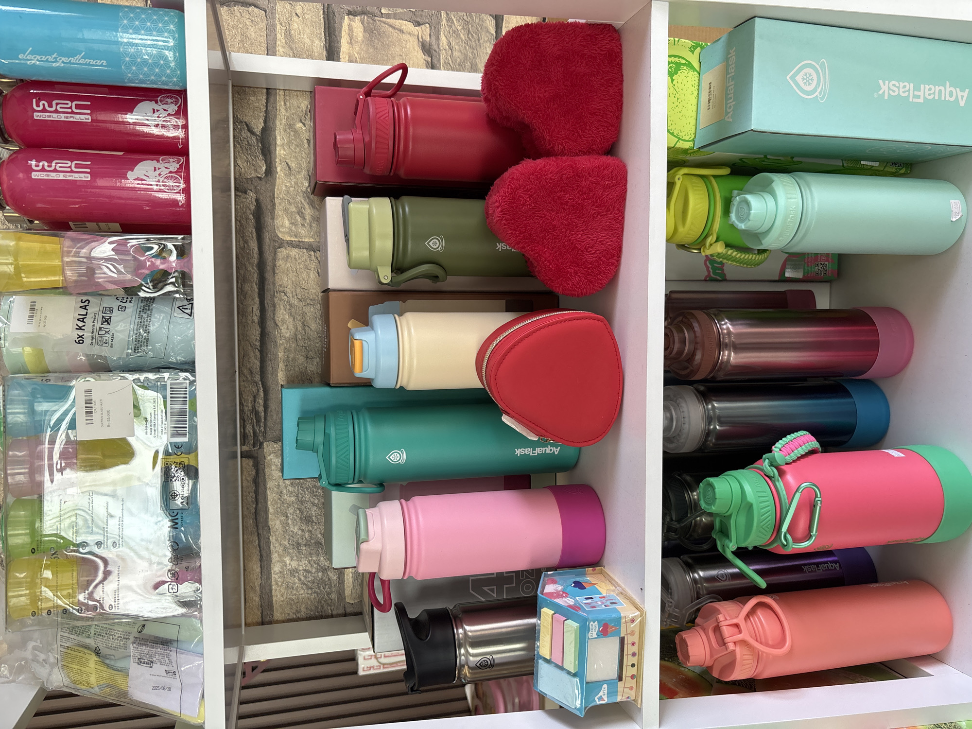 The image displays a shelving unit filled with various water bottles and related items. The shelves are white, and the background behind them appears to be a stone or brick wall.
On the top shelf, several packages of reusable cups are visible, some with colorful contents. There are also several tall, slender water bottles in bright pink, light blue, and one with a "WRC" logo repeated.
The middle shelf is densely packed with more water bottles of different sizes and colors, including pink, teal, cream, olive green, and red. Some have prominent brand names like "AquaFlask" printed on them. A red, heart-shaped pouch and two fluffy red heart-shaped objects are also present on this shelf. A small, decorative house-shaped object, possibly a dispenser or toy, sits on the far left.
The bottom shelf features more water bottles in various shades of pink, teal, and metallic silver. Some bottles have attached carabiners or straps. A light blue box with the "AquaFlask" logo is visible on the right side of this shelf.
The overall impression is of a retail display or a personal collection of reusable drinkware. The lighting suggests an indoor setting. There are no people visible in the image. The location of Gajahan, Indonesia, is not directly evident from the visual cues within the image itself.
