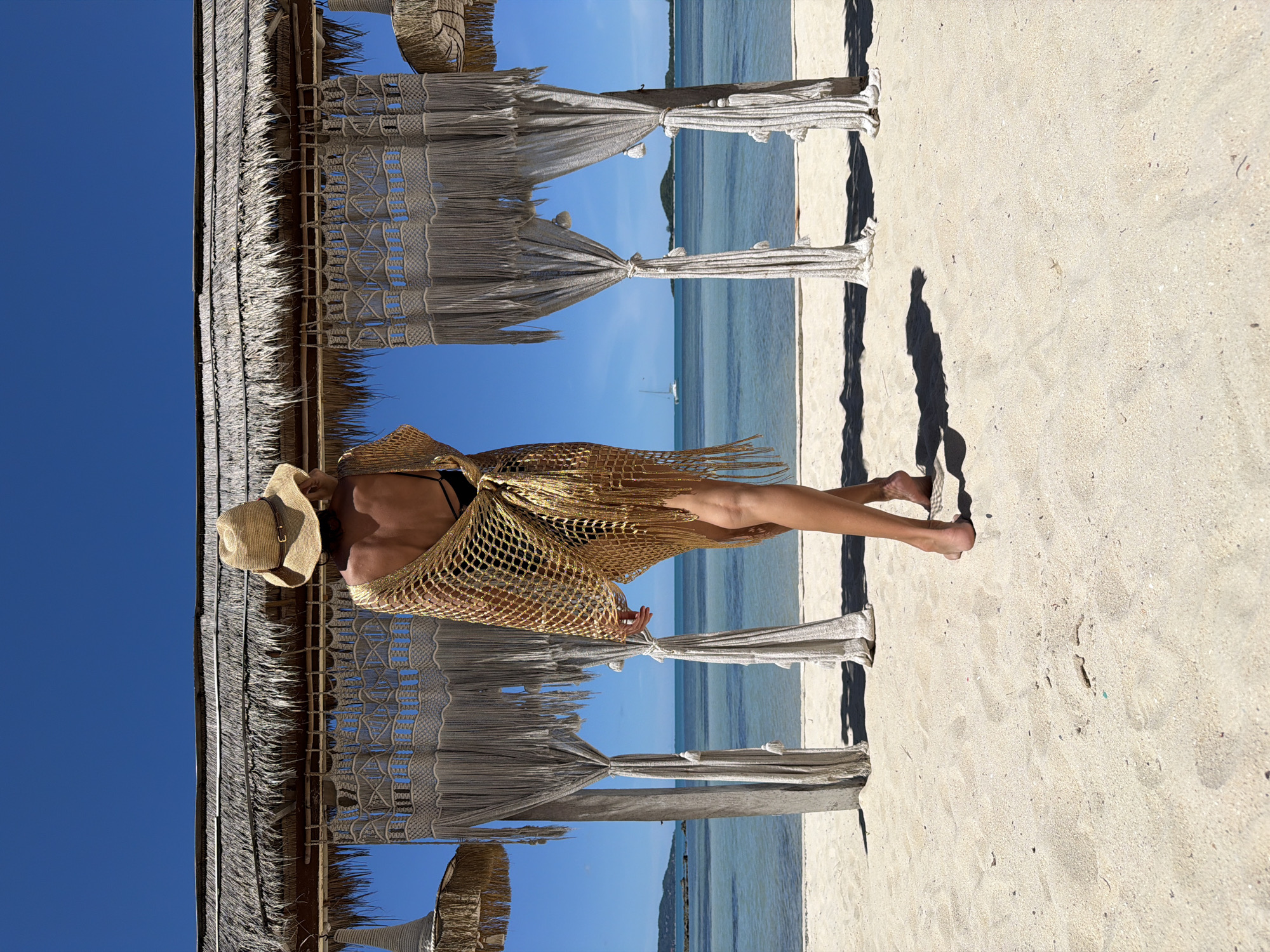 A woman wearing a straw hat and a gold net cover-up stands on a sandy beach in Baan Thong Krut, Thailand. She is facing away from the camera, looking out at the ocean. Behind her is a bamboo structure with a thatched roof and macrame curtains. The ocean is calm and blue, with a sailboat visible in the distance. The sky is clear and blue. The sand is light in color and appears to be fine-grained.