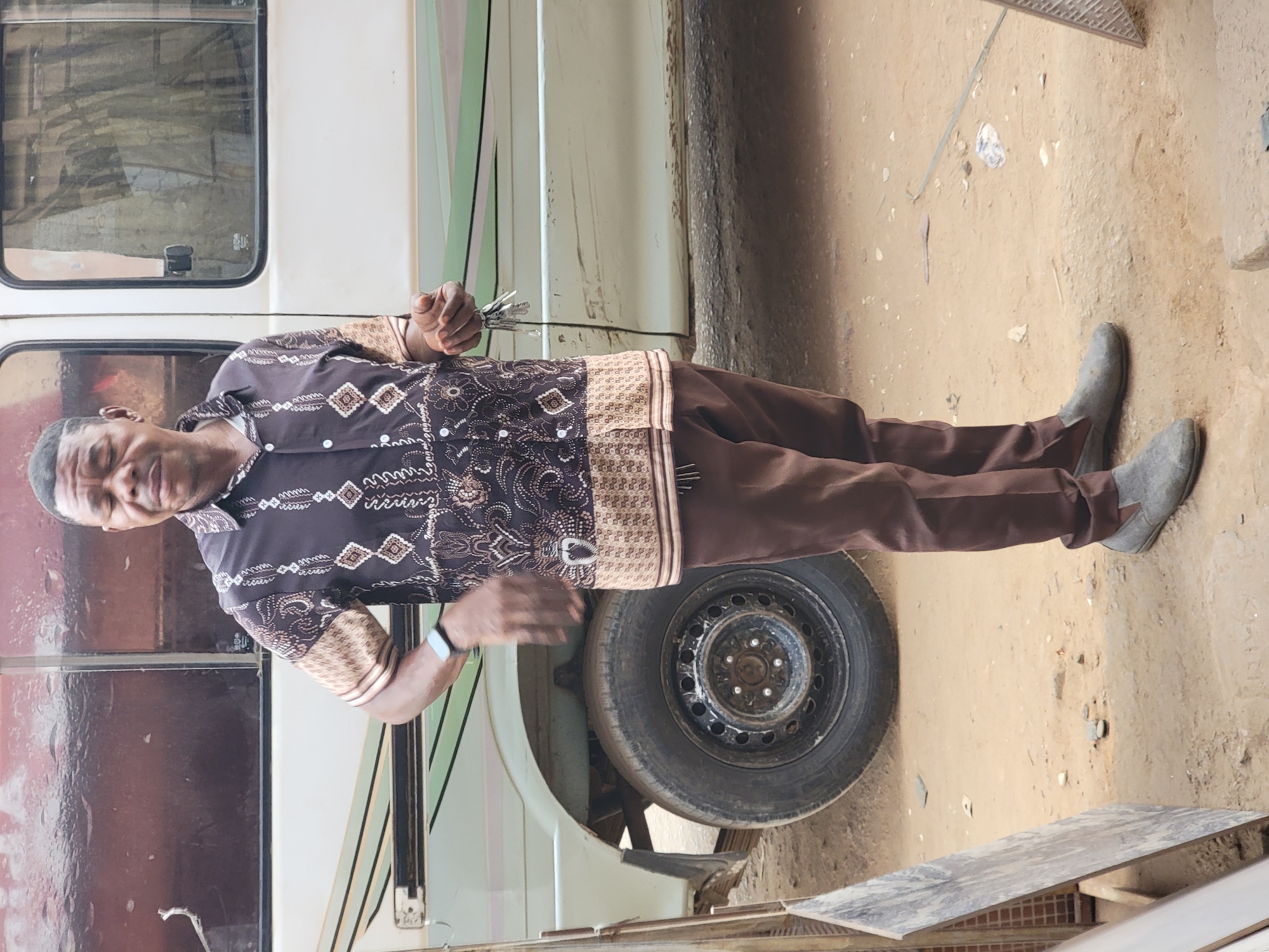 An African man stands outside, holding keys in his right hand and gesturing with his left. He wears a patterned button-down shirt and brown pants, with gray shoes. Beside him is the side of a van and a tire. The ground is dirt. Location: Maiburiji, Nigeria.