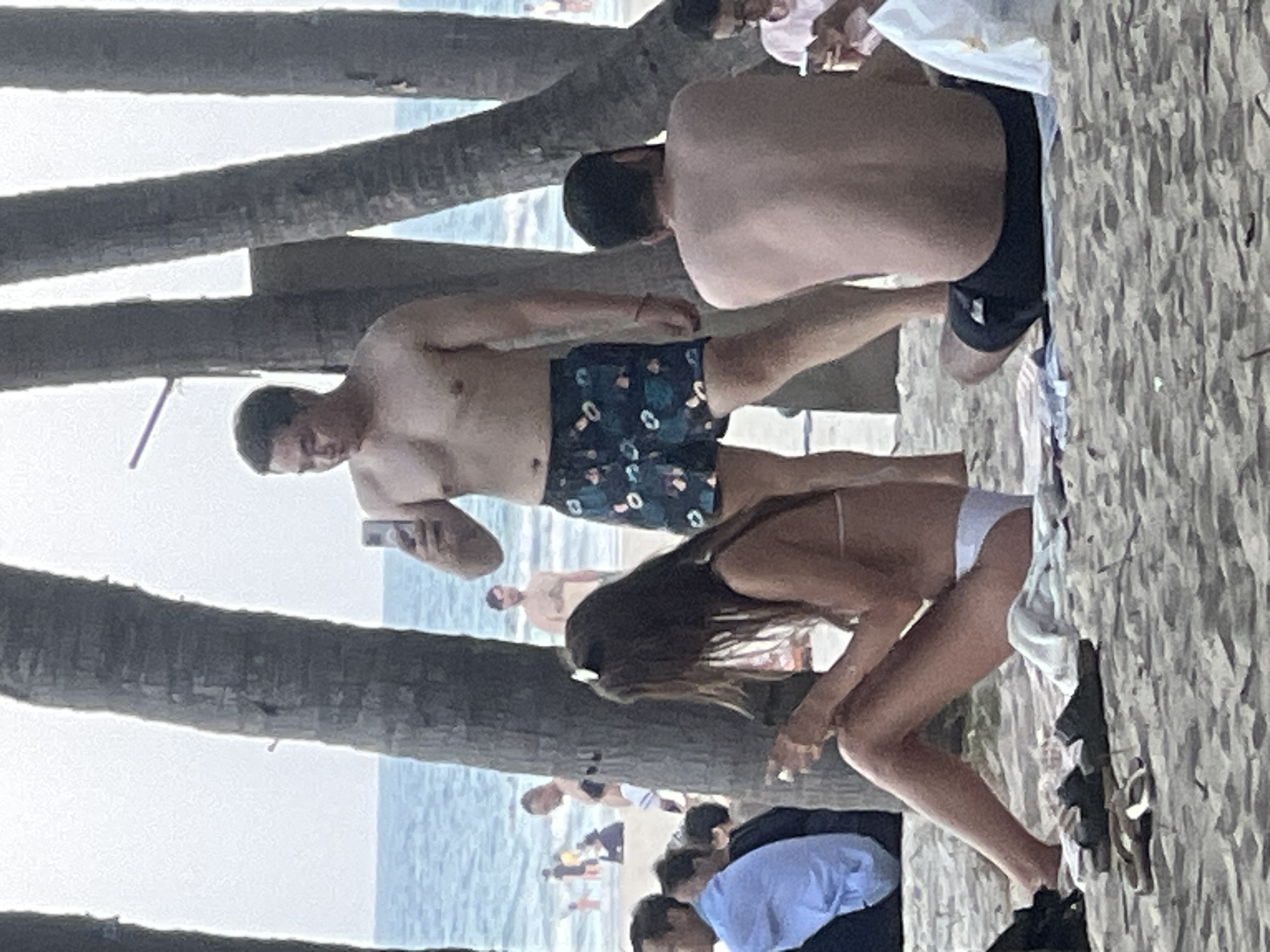 This image, taken on a beach in Da Nang, Vietnam, depicts a group of people enjoying a day by the ocean. In the foreground, a woman with long brown hair sits on the sand wearing a white bikini, holding a smartphone. Next to her are a pair of black sandals and a pair of flip-flops. Behind her, a man stands shirtless, wearing patterned swim trunks, and holds a smartphone, possibly taking a selfie. Further in the background, several other individuals are visible on the beach and in the water. The scene is characterized by the presence of palm trees, the sandy shore, and the blue ocean under an overcast sky.