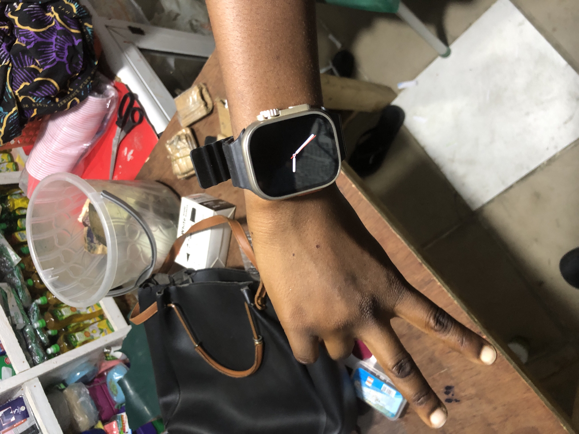 The image shows a person's arm and hand, with a smartwatch on the wrist, in what appears to be a stall or shop. The background is cluttered with various items, including bottles of liquid on shelves, a large transparent bucket, pink disposable cups, scissors, fabric with a purple floral pattern, and what looks like packaged goods. The table surface is made of wood, and a black handbag with a tan strap is visible. The person is making a peace sign with their fingers. The scene suggests a commercial setting, possibly in Ado, Nigeria, given the context provided, where goods are displayed and sold.