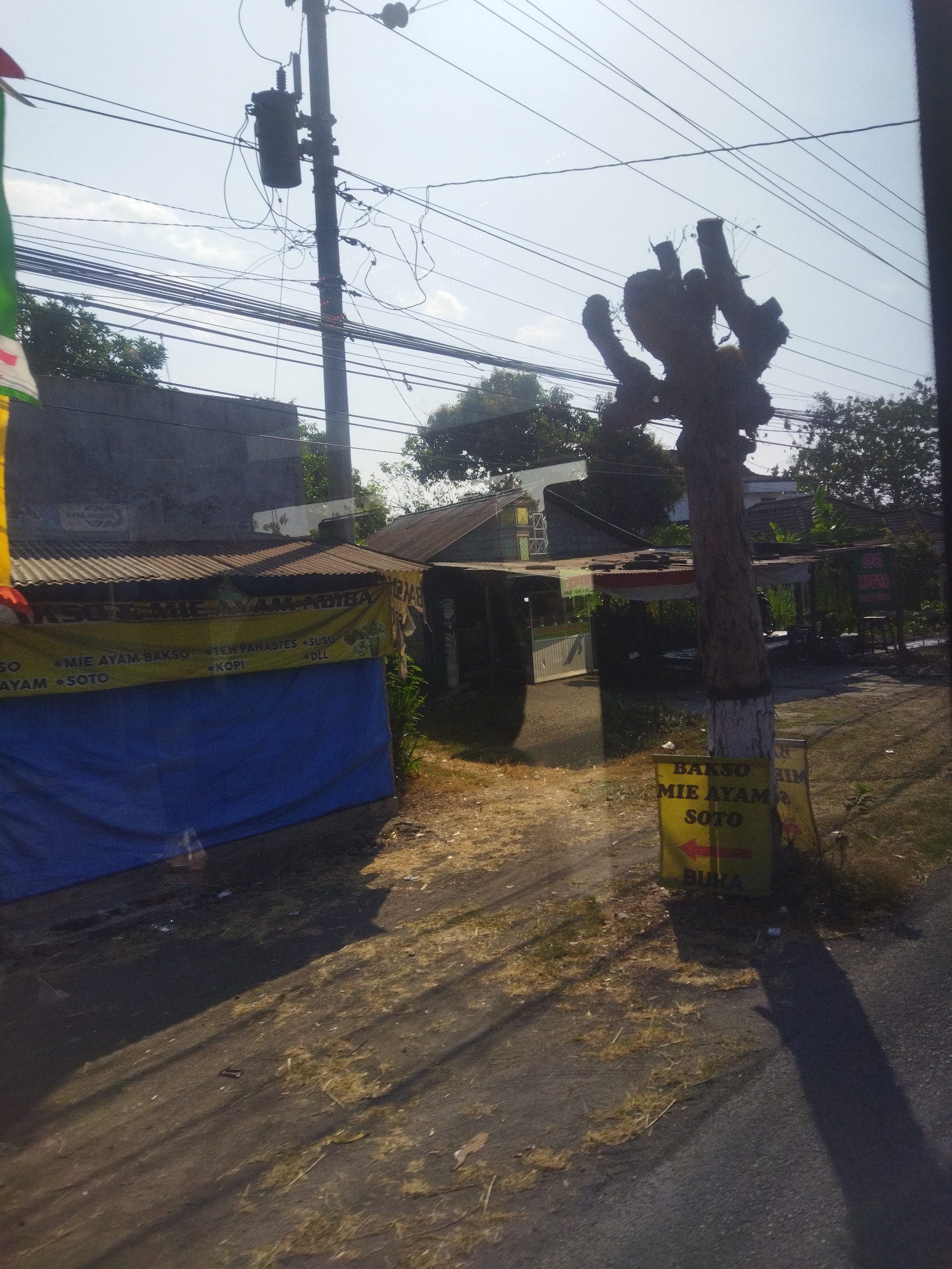 The image captures an outdoor scene in Daleman, Indonesia, characterized by a mixture of urban infrastructure and vegetation. A prominent electrical pole with a transformer and numerous wires dominates the upper left portion of the frame. In the foreground, a dirt path leads towards a small building with a corrugated metal roof, possibly a food stall or shop, identified by a yellow banner displaying text in Indonesian, including "MIE AYAM ADIBA," "MIE AYAM BAKSO," and "SOTO." A blue tarp is partially visible in front of the structure. To the right, a peculiar, pruned tree stands, its branches bare and gnarled. A yellow sign with red text, also in Indonesian, is propped up near the tree, indicating "BAKSO," "MIE AYAM," and "SOTO" with an arrow pointing left. The overall environment suggests a roadside setting with evidence of commerce and the presence of electrical utilities. The sky is clear and bright, indicating daylight.