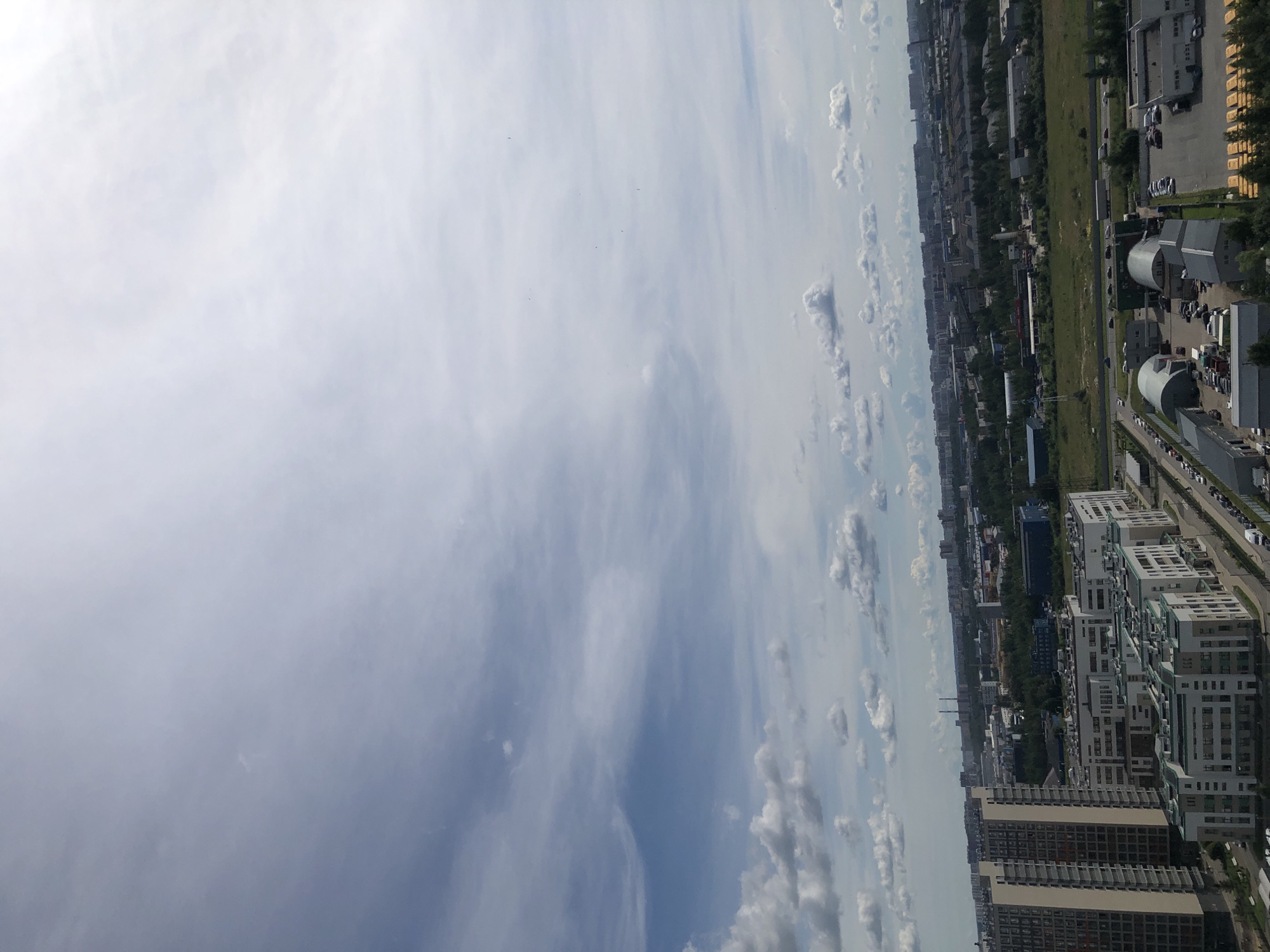 This media file captures an aerial view of a suburban or urban landscape on a clear day. The scene is dominated by a vast expanse of sky filled with scattered clouds, ranging from wispy cirrus formations to more dense cumulus clouds. Below the sky, the landscape unfolds, revealing a mix of residential buildings, industrial structures, and patches of greenery.
The residential buildings appear to be modern, with multiple stories and varied architectural styles, suggesting a developed area. Some are tall apartment complexes, while others are lower-rise structures. Industrial buildings, characterized by their functional design, are also visible, alongside roads with a few cars driving. A long, green strip of land, possibly a park or undeveloped area, runs through the scene, interspersed with trees.
The image appears to be taken from a high vantage point, possibly a drone or a tall building, looking down and to the side. The lighting suggests it is daytime, with the sun likely high in the sky, casting soft shadows. The weather seems pleasant, with the presence of clouds indicating a dynamic sky rather than a completely overcast or clear blue one. No people are directly visible in the image, and there is no discernible text. The overall impression is one of a developing or established residential and industrial area, with a clear sky and daytime activity.