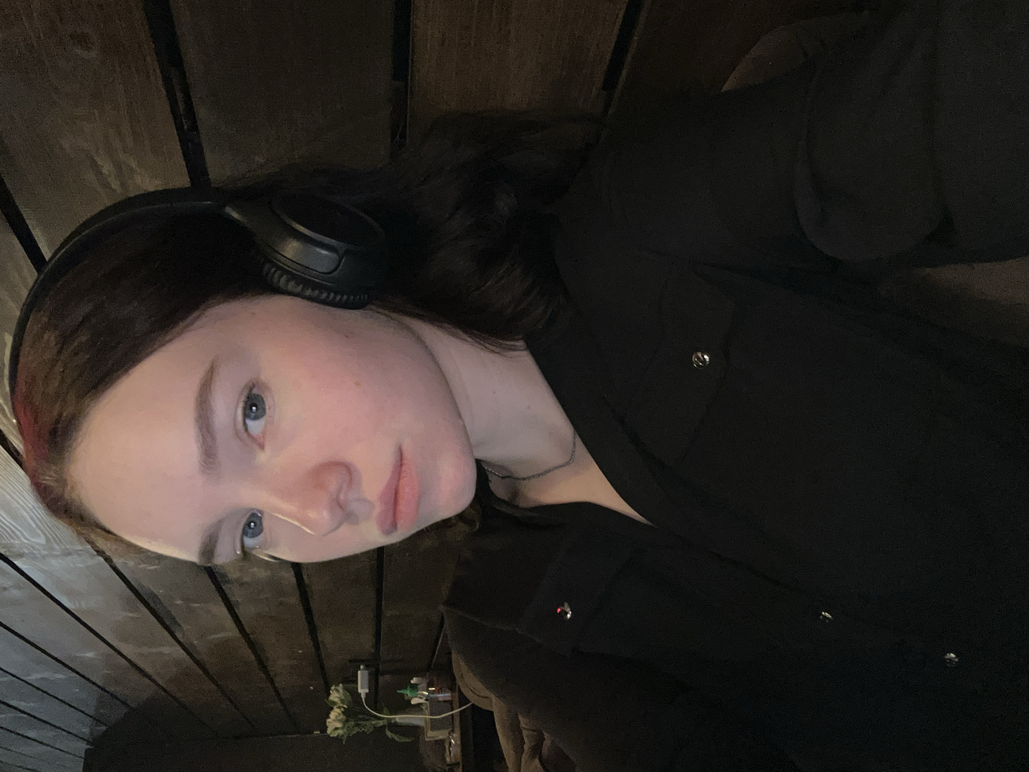 A young woman with fair skin, blue eyes, and dark hair wearing black headphones and a black button-down shirt poses for a selfie. She appears to be indoors, seated near a wooden wall or surface. A small bouquet of white flowers and miscellaneous items are visible in the background. There is no verifiable information indicating the image was taken in Chaikovsky, Russia.