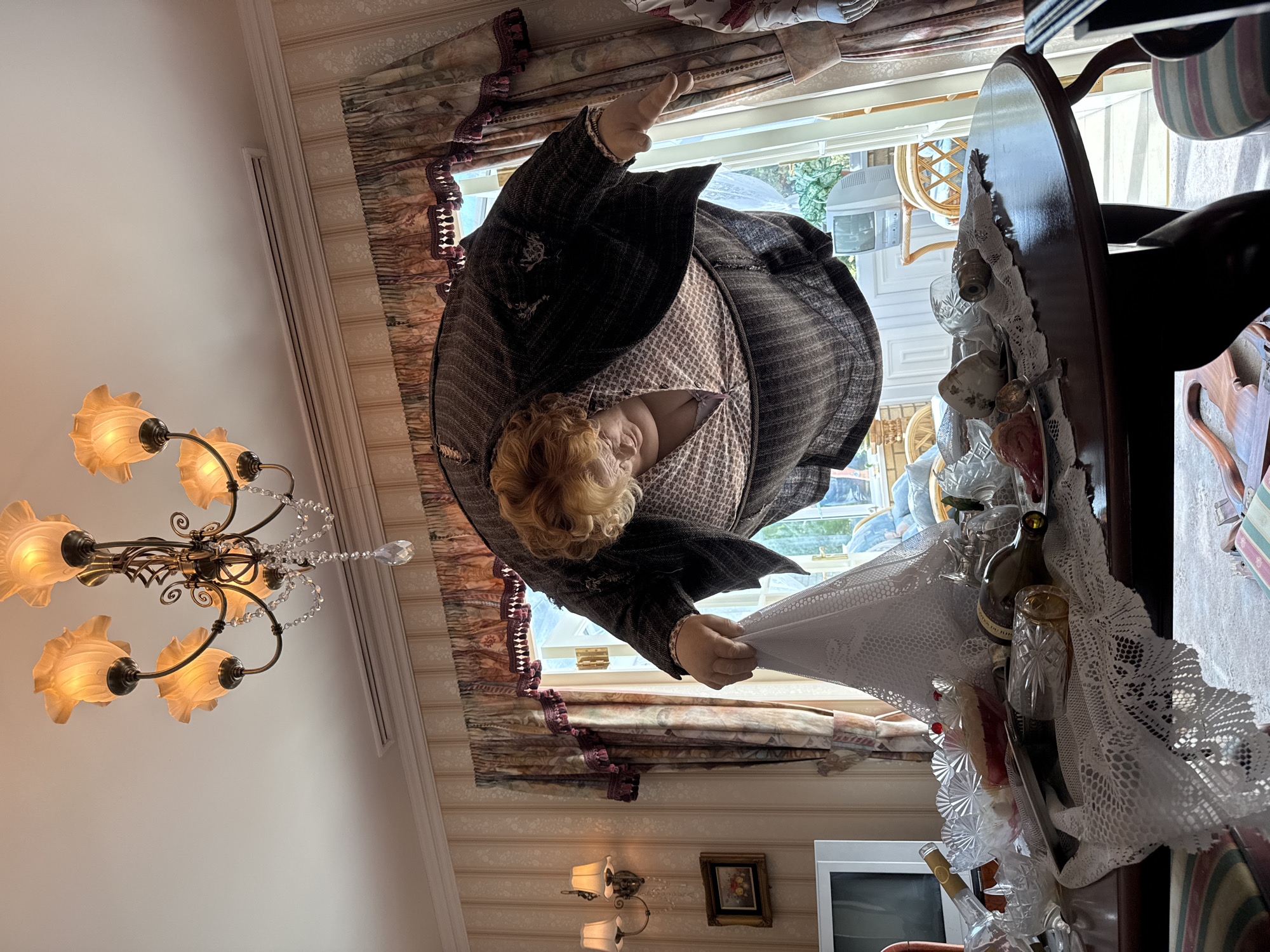 A rotated indoor scene features a wax figure dressed in a tweed suit with a floral blouse, seemingly displaying a white lace fabric. The figure is positioned near a window with floral curtains, overlooking what appears to be an outdoor scene with a white wicker chair and glimpses of greenery. To the right, a dark wooden table or cabinet is adorned with various glassware, bottles, and lace trim. A chandelier with floral shades hangs from the ceiling on the left. In the background, a white wall and television are visible, along with a framed picture and a wall lamp. The location, Nerima, Japan, is inferred from context not directly visible in the image.