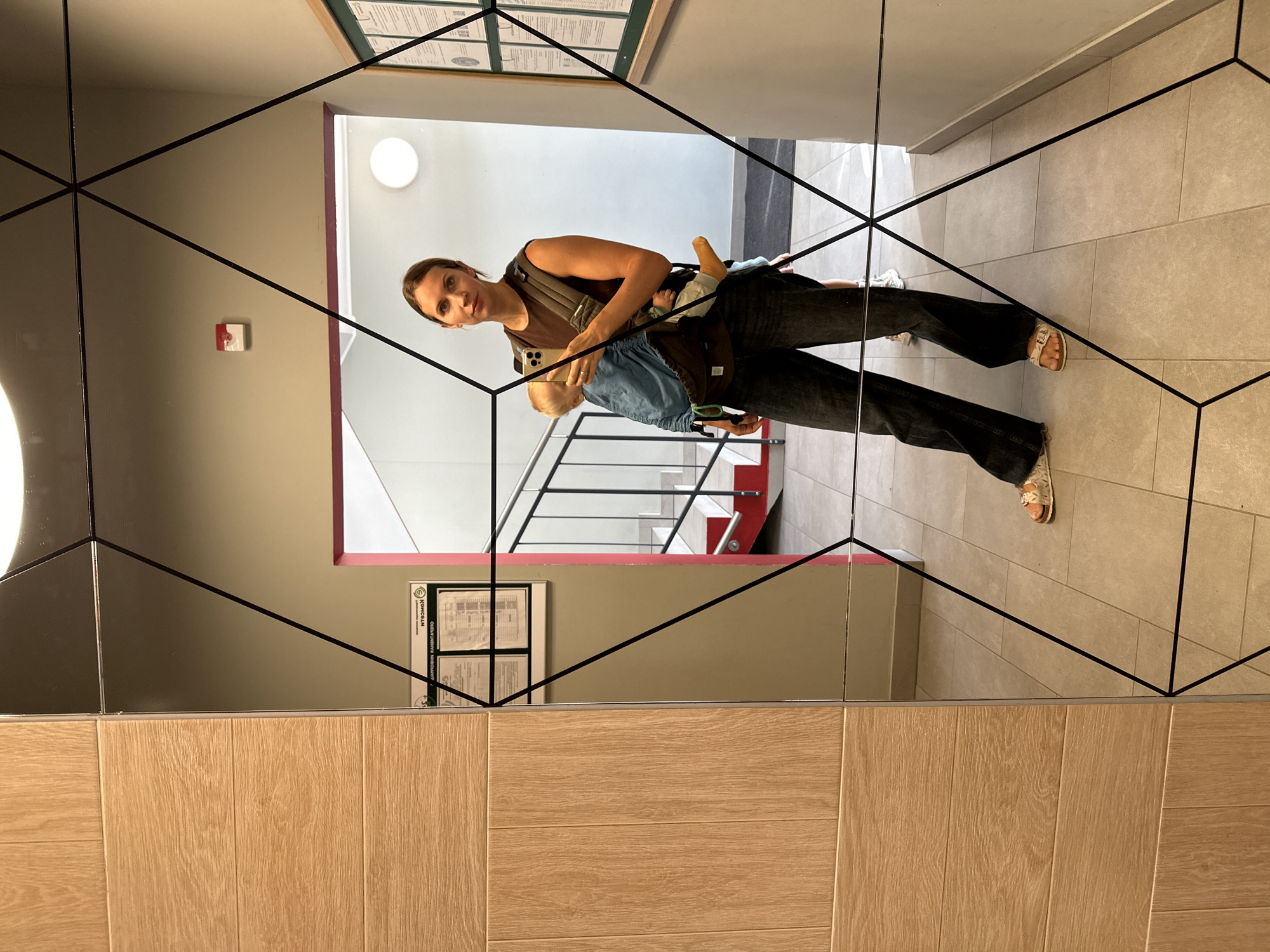 A woman is captured in a mirror's reflection, standing in what appears to be a well-lit, modern indoor space, possibly a building lobby or hallway. She is holding a baby in a carrier attached to her chest. The woman is looking directly at the camera, holding her phone to take the selfie. Her attire consists of a dark, sleeveless top, dark, wide-leg jeans, and light-colored sandals. The baby is wearing a light blue carrier and has a yellow sock on its right foot.
The environment shows a wall with wood-paneled sections on the left, transitioning to a large, geometric-patterned mirror that dominates the reflection. In the background, a staircase with red accents and metal railings is visible. A bulletin board with documents is mounted on the wall to the right. The floor is tiled with light-colored square tiles. The overall lighting suggests it is daytime. The location is identified as Vsevolozhsk, Russia.