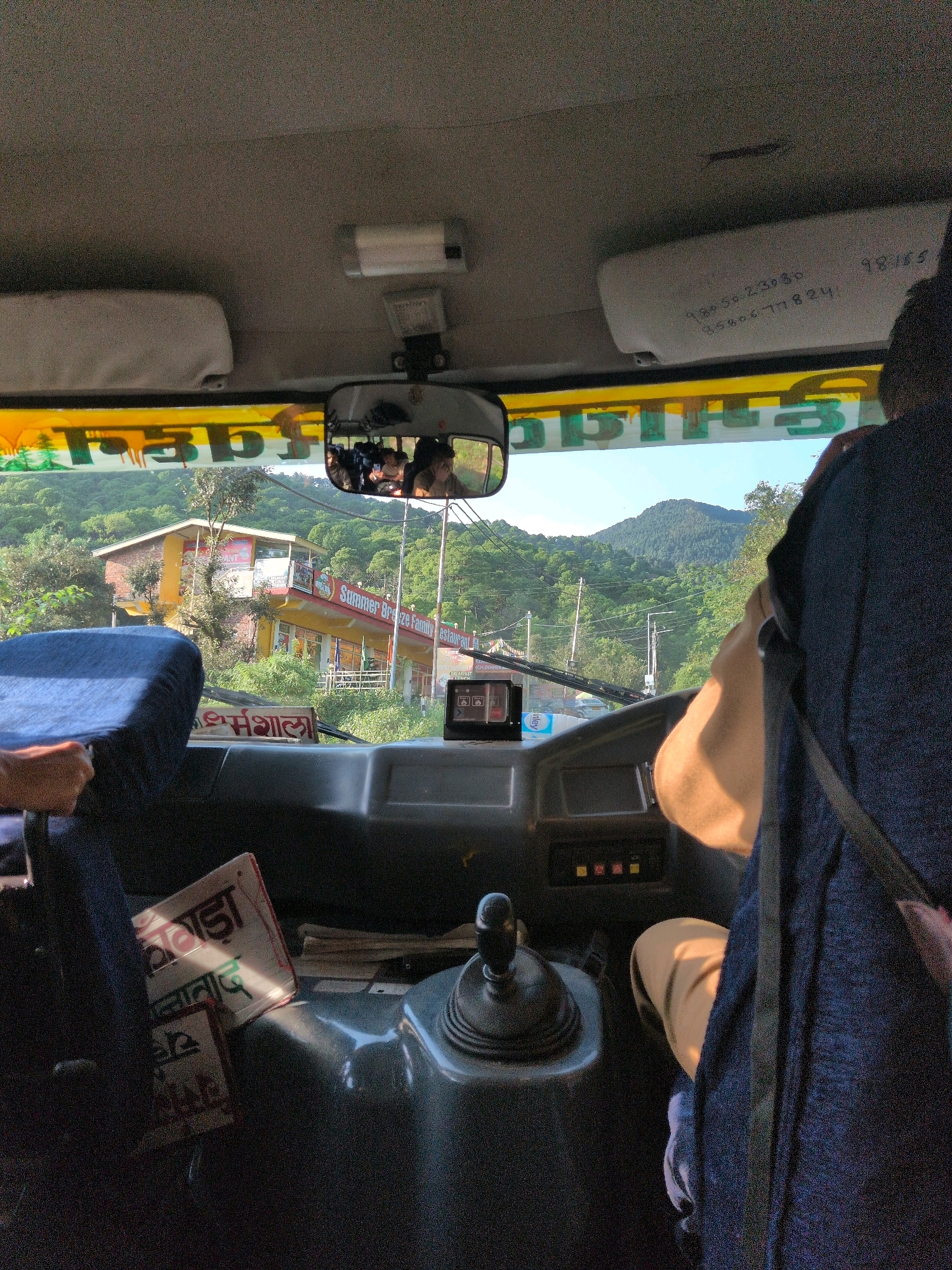 The image shows the interior of a bus, looking forward through the windshield and past the driver. The driver, wearing a dark blue shawl or jacket and tan pants, is visible from the back and right side. The bus is on a road in a hilly, green environment, likely in or near Dharamshala, India. A building with a yellow facade and red trim, identified as "Summer Breeze Family Restaurant," is visible on the right side of the road. There are trees and mountains in the background, with a clear blue sky.
Inside the bus, a rear-view mirror reflects a number of passengers. A sun visor on the ceiling has handwritten text and numbers. On the dashboard, a small electronic device is mounted. The gear stick is prominent in the center console. A blue fabric seat is visible on the left. A book or pamphlet with text in a non-Latin script is on the dashboard, partially illuminated by sunlight.