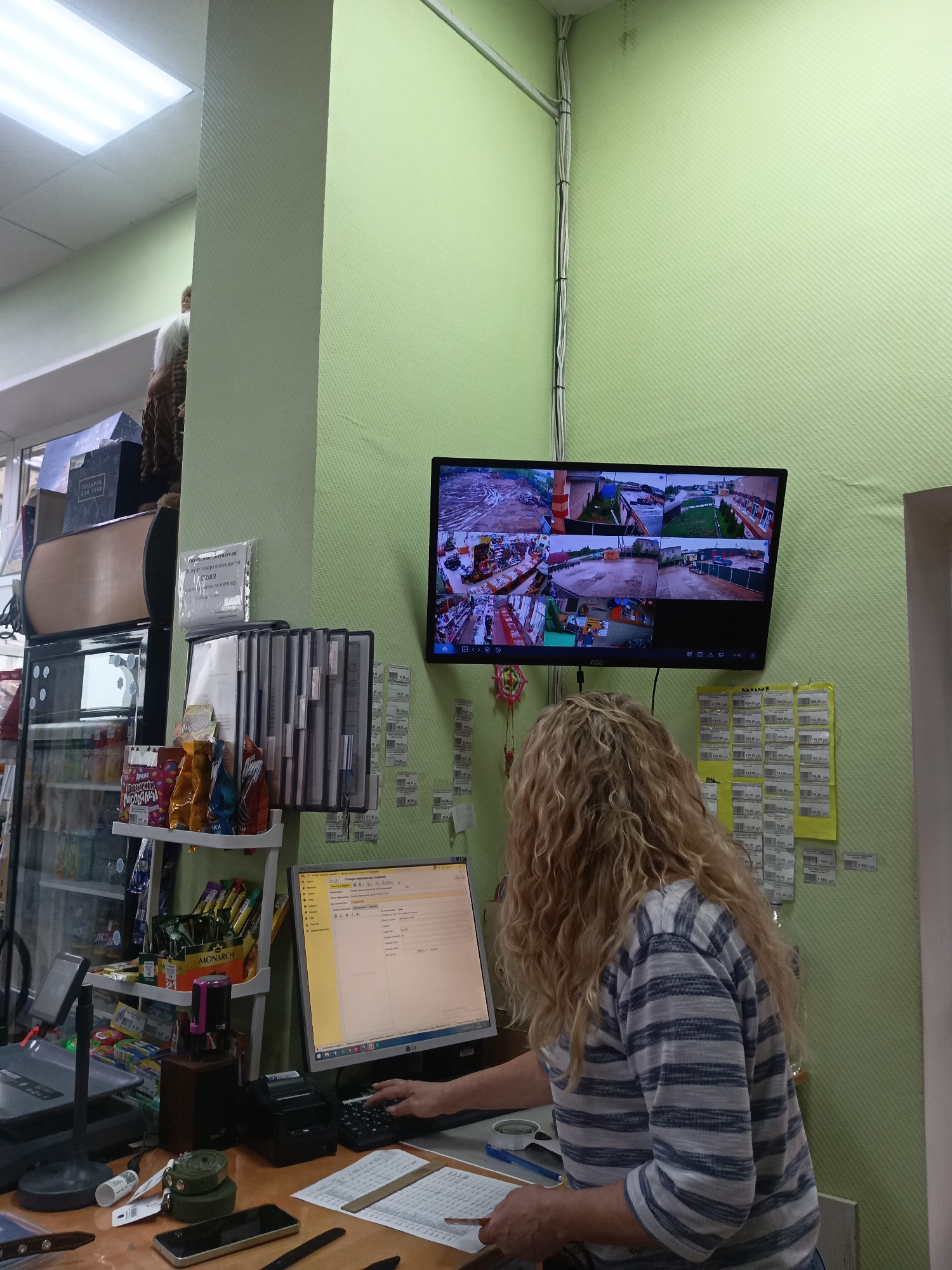 The image shows a woman with blonde, curly hair standing at a counter in a retail environment, possibly a shop or small store. She is wearing a blue and white striped shirt and is facing a computer monitor that displays what appears to be an application. A security monitor is mounted on the light green wall, displaying feeds from multiple security cameras showing an industrial/construction area, potentially a shipping dock or yard, likely related to the business. Various items such as snacks, papers, a calculator, and a telephone are on the counter. Documents or price lists are affixed to the wall near the security monitor. Location: Фоки, Russia.