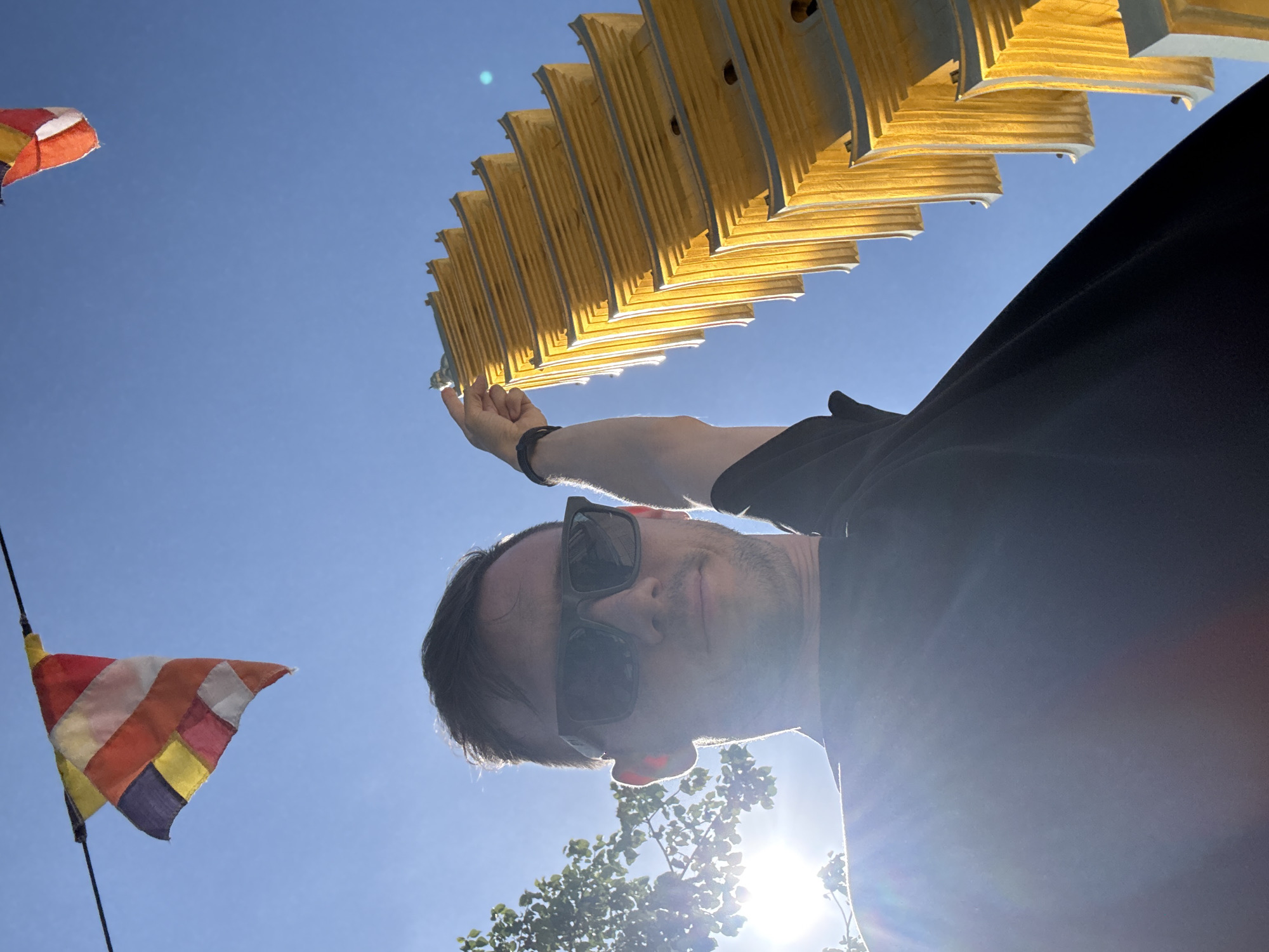 A man in sunglasses and a black t-shirt is holding a folded yellow object over his head, as viewed from a low angle against a bright blue sky. The man is partially obscured by the light. A Buddhist flag is visible in the lower left of the frame. Sunlight is causing lens flare. The location is Diên Khánh, Vietnam.