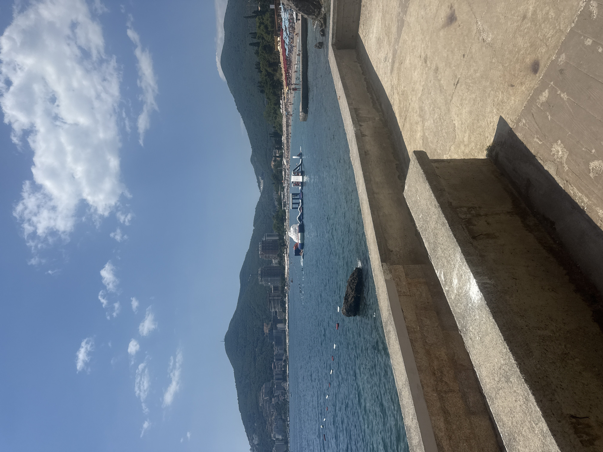 The image depicts a sunny day at a beach in Budva, Montenegro. The foreground shows a concrete walkway with a low, weathered concrete wall. Beyond the wall, the blue sea is visible, with a few buoys marking a swimming area. In the water, there's an inflatable water park. The middle ground features a crowded beach with numerous umbrellas and sunbathers. Buildings, likely hotels and apartments, are clustered on the hills in the background. Two large, green, tree-covered mountains dominate the skyline. The sky is a clear blue with scattered white clouds. The overall impression is a lively summer scene at a popular coastal resort.