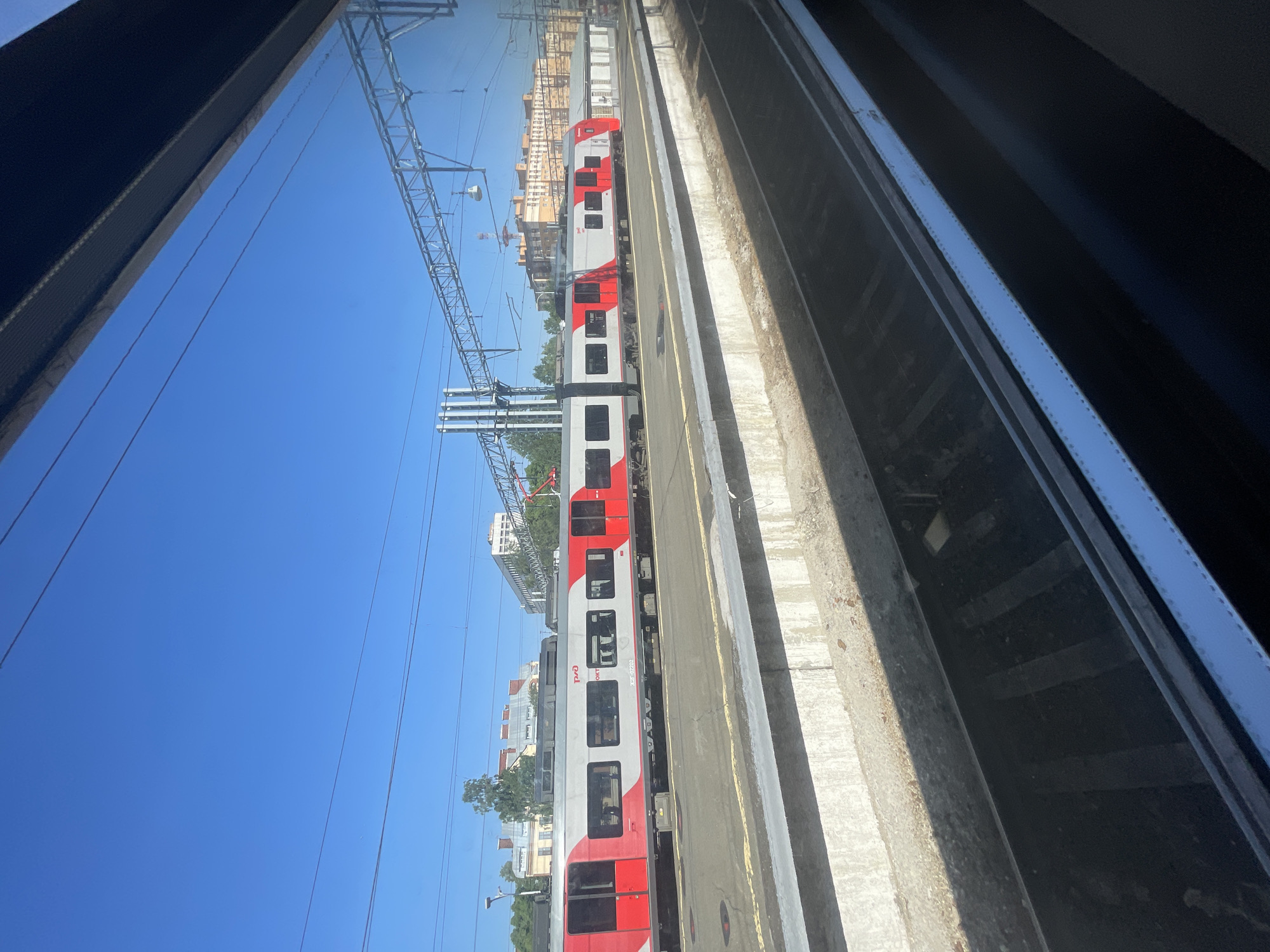 A vertically oriented image, captured from the window of another train or an elevated platform, displays a modern red and white electric train stationary on a railway track next to a concrete platform. The sky above is a clear, vibrant blue, indicating bright daylight and fair weather.
The train, a multi-car unit, features a distinctive livery of white with a bold red stripe running along its lower half and around the doors. Several large windows are visible along its side. Prominent text on the train includes the "РЖД" (Russian Railways) logo and the designation "ЭГ2Тв 020", identifying it as a Lastochka (Swallow) train.
Overhead, a complex network of metal gantries and power lines (catenary system) stretches across the blue sky, signifying an electrified railway. In the background, beyond the train and some trees, stand multiple-story buildings, characteristic of an urban or suburban area. The immediate foreground on the right shows a dark, shadowed section of railway infrastructure or the window frame from which the picture was taken. The scene captures a typical railway environment in Saint Petersburg, Russia.