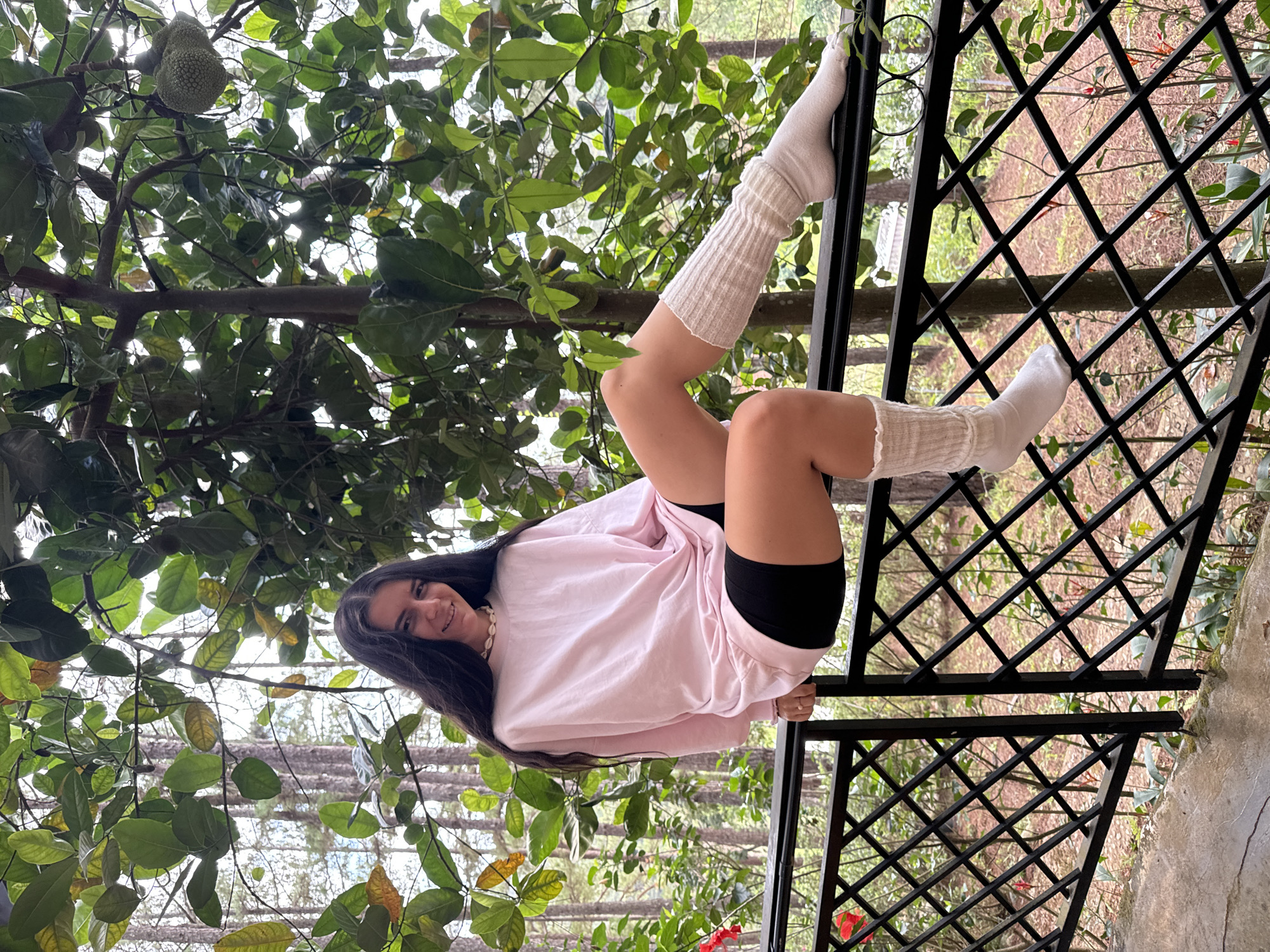 A young woman with long, dark hair is perched on a black, diamond-patterned metal railing. She wears a light pink long-sleeve shirt, black shorts, and white, textured, calf-length socks. Her legs are crossed and extended to the upper left of the frame, with her feet resting on the railing. The background features lush green foliage, including a tree with large, green fruits hanging from its branches. Beyond the foliage, trees are visible. The location is Da Lat, Vietnam.
