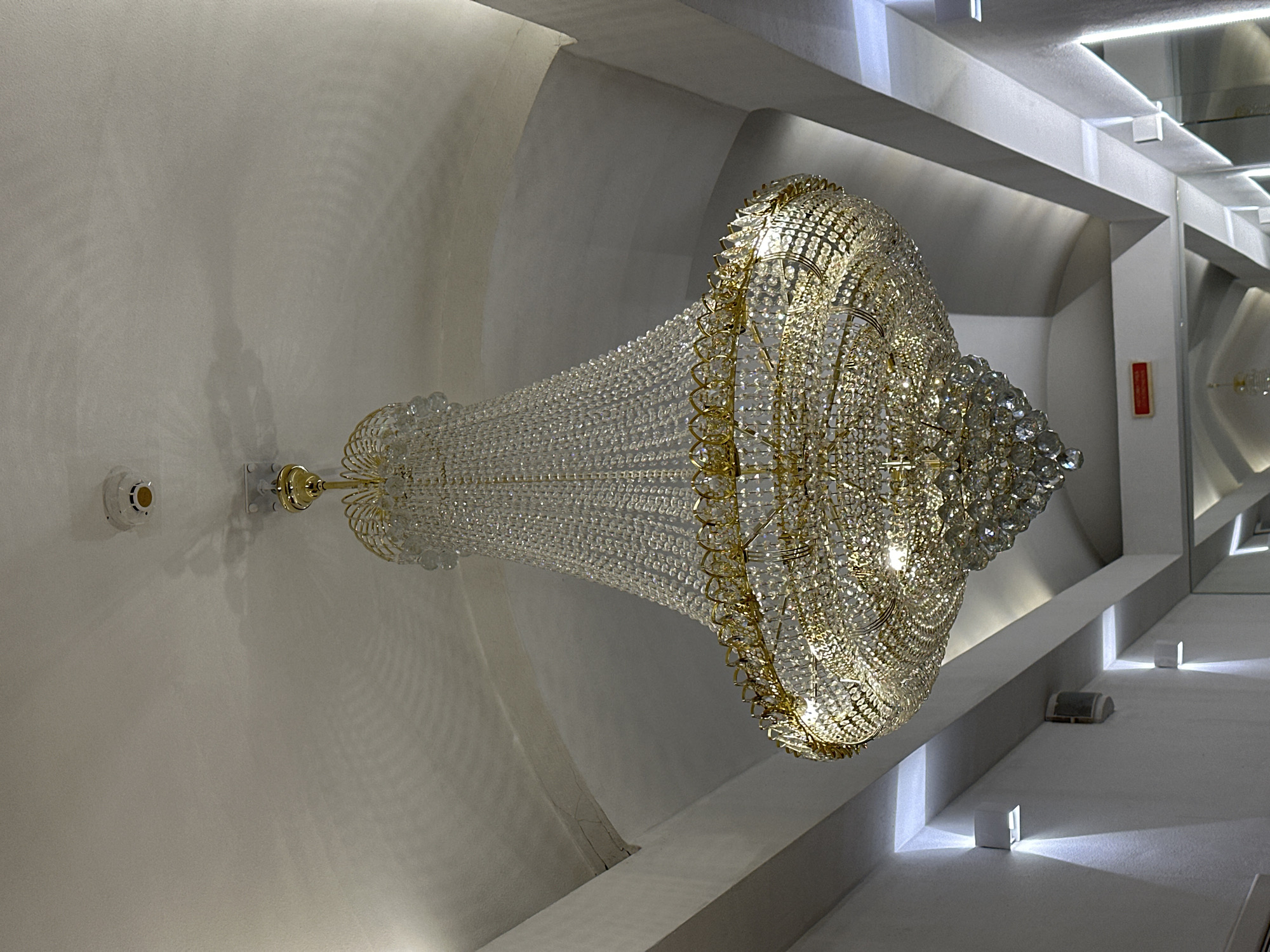 The image displays a large, ornate chandelier suspended from a white ceiling in what appears to be a luxurious interior space, specifically in Moscow, Russia. The chandelier is predominantly made of numerous clear crystal beads and drops, intricately arranged on a golden metallic frame. It is brightly illuminated, casting a warm, sparkling light and intricate patterns onto the surrounding surfaces.
The ceiling is a smooth, off-white surface, featuring a modern architectural design with recessed panels and linear lighting elements. These recessed sections create a sense of depth and provide additional ambient illumination, enhancing the overall bright and clean aesthetic of the space. To the upper left of the chandelier's mounting point, a white smoke detector or similar alarm device is visible on the ceiling.
Along the right side of the image, the wall features multiple white, cube-shaped light fixtures that emit focused beams of light upwards and downwards, adding to the contemporary lighting scheme. In one of the recessed wall sections further back, a small, red rectangular sign with white text is visible, likely an emergency or fire safety indicator, though the text itself is not clearly decipherable.
Faint shadows, possibly of a person or objects not directly in view, are cast on the light-colored surfaces, suggesting the presence of other elements or the photographer's shadow. The overall scene depicts a grand and modern interior, likely a lobby, hallway, or event space, with a focus on sophisticated lighting and elegant design. The time of day is not determinable, as the interior is lit by artificial light.