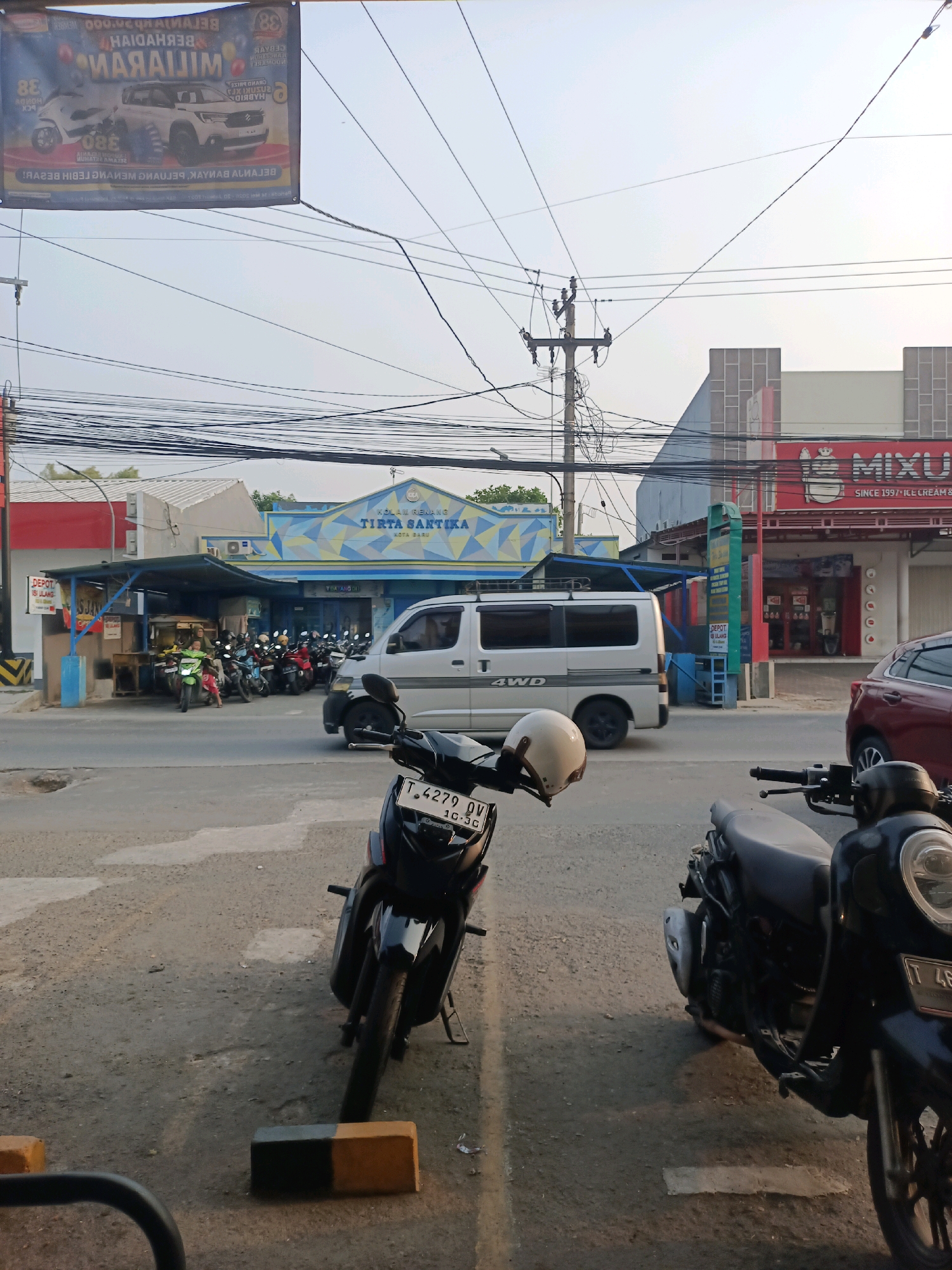 The image captures a street scene in Karawang, Indonesia, with a clear view of a building featuring a sign that reads "TIRTA SANTIKA KOTA BARU". The building has a distinctive blue and yellow patterned facade. In front of the building, a row of parked motorcycles is visible, with a green motorcycle in the foreground being ridden by an individual whose legs are visible. To the right of the building, another establishment with a red sign displaying "MIXU" is present, and it appears to be an ice cream shop. A white van with "4WD" written on its side is also in motion on the street. The foreground of the image is occupied by two parked motorcycles, one black and the other a darker shade. Power lines crisscross the sky above the street, and a utility pole is also visible. The lighting suggests it is daytime, possibly late afternoon, given the shadows.