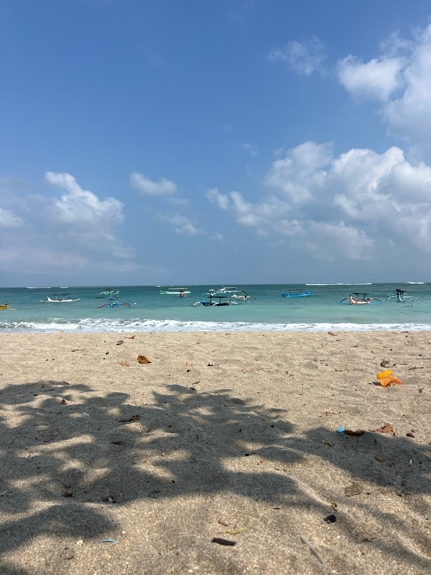 An outdoor, eye-level shot captures a serene beach scene on a clear day. The foreground is dominated by sand, with a distinct shadow pattern cast by unseen foliage, suggesting a nearby tree. A few dry leaves are scattered across the sand, along with some small debris. A bright orange object, possibly a piece of plastic, is visible on the right side of the frame.
The middle ground features the gentle turquoise water of the ocean, with small waves lapping at the shore. Numerous traditional outrigger boats, known as jukungs, are moored in the shallow water. Some are empty, while others appear to have people on board, casually relaxing. The boats vary in color, with prominent blue and white designs.
The horizon line is sharply defined, separating the ocean from the sky. The sky itself is a vibrant blue, dotted with fluffy white clouds. The overall impression is one of tranquility and a typical coastal landscape in Kuta, Indonesia, a popular tourist destination known for its beaches and traditional boats.