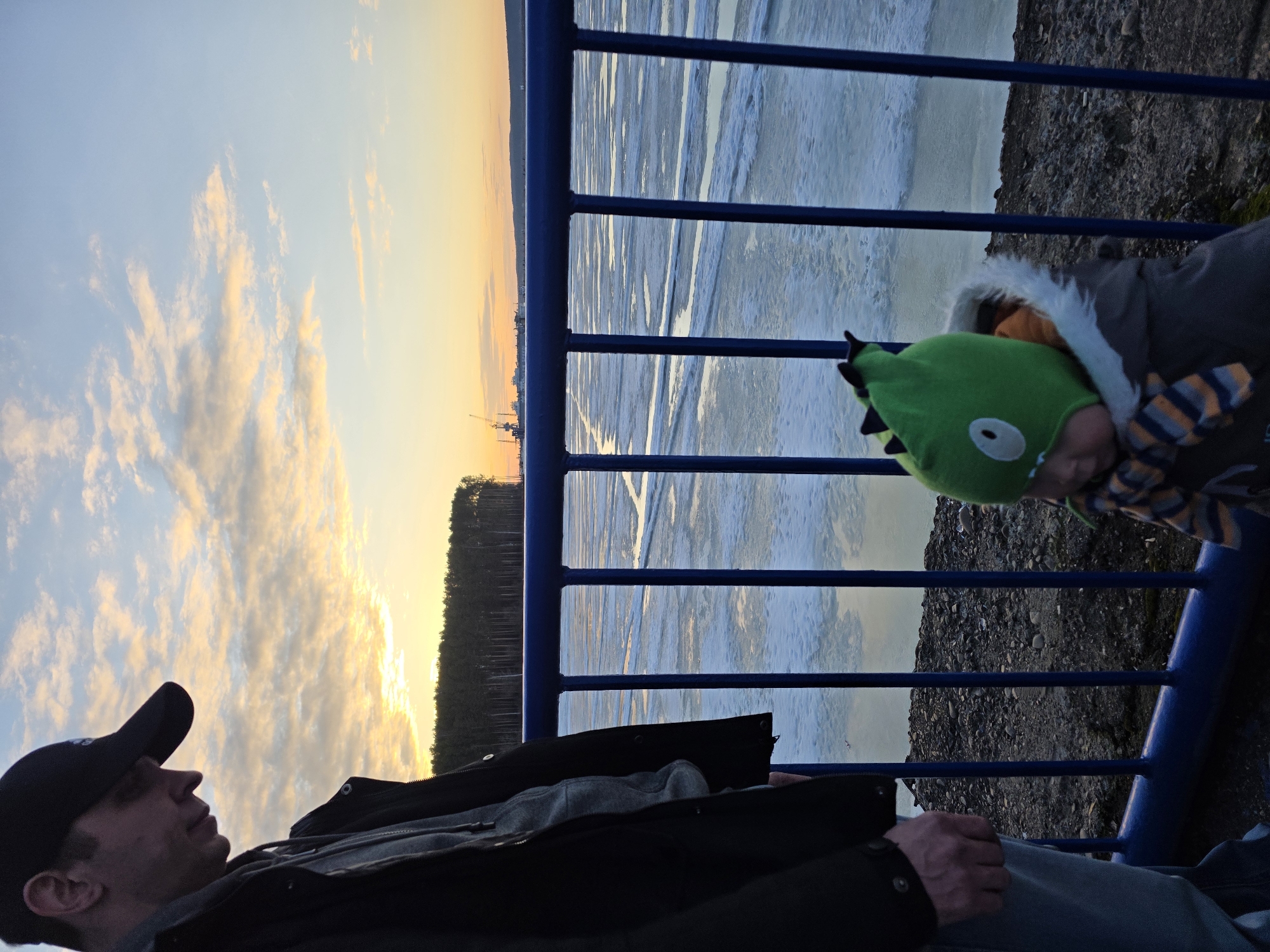 The image depicts two individuals beside a blue metal railing, with a partly frozen body of water visible beyond. The person in the foreground is wearing a black baseball cap, a dark jacket, and jeans. The second person, likely a child, is wearing a green dinosaur-themed hat and a warm coat. The background includes a line of trees and a colorful sunset sky. The location is near the city of Chaikovsky in Russia, as indicated by the presence of frozen bodies of water and architecture. The image is rotated 90 degrees counter-clockwise.