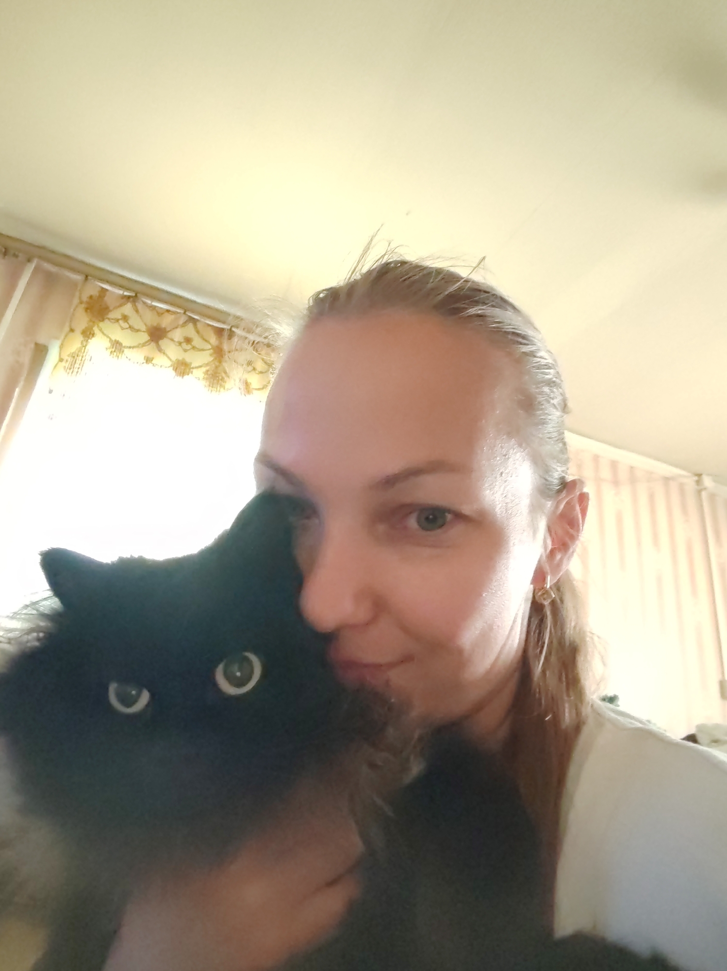 The image is a close-up selfie of a woman with a black cat. The woman is looking towards the camera with a neutral expression, and her face is partially obscured by the cat. The cat, also black, is positioned in front of her, with its eyes visible and looking towards the camera. The background appears to be an indoor setting with curtains and striped wallpaper. The lighting is bright and seems to be coming from a window. The image quality is somewhat blurry, likely due to the close proximity of the subjects and the camera. This image has been geotagged to Чухлэм, Russia.