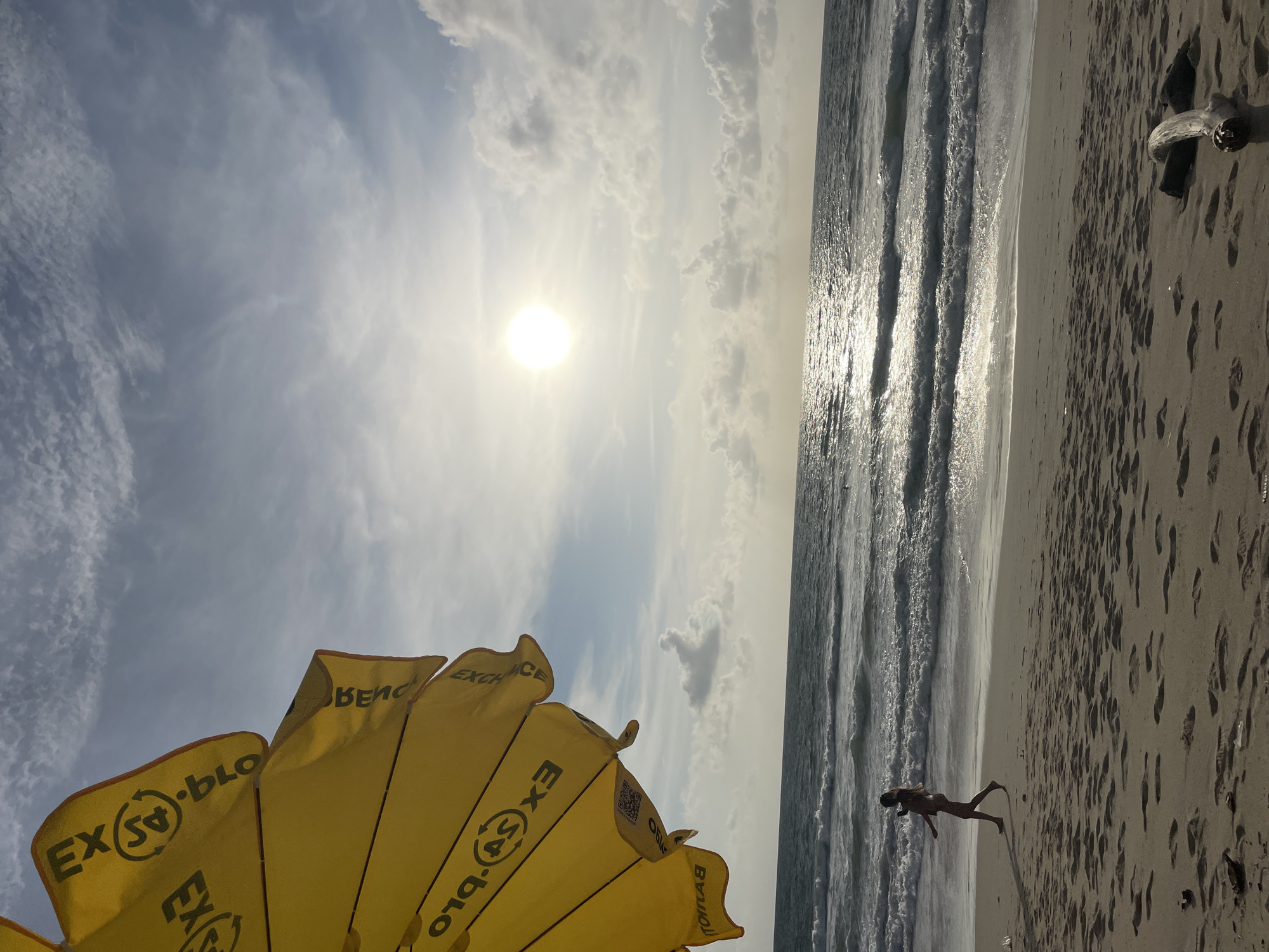 This photo shows a beach scene in Choeng Thale, Thailand. The view is oriented vertically with the horizon on the right. In the foreground is the lower portion of a yellow beach umbrella with the text "EX.BLO EXCHANGE." The background shows a sunny sky with scattered clouds and the ocean. A thin strip of sand runs along the right edge of the image. A single person is visible on the sand, near the water's edge. There is a piece of driftwood on the sand.