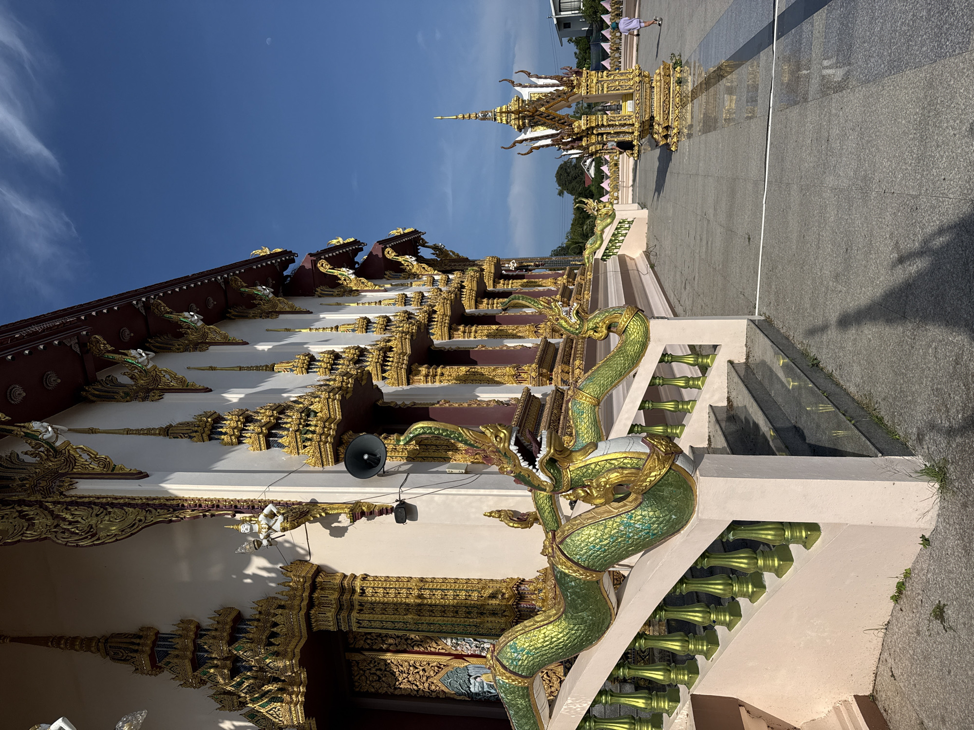 This is a low-angle, full outdoor shot of a Buddhist temple in Ko Samui District, Thailand. The temple is ornate, with gold and deep red trim, and features intricate carvings and statues, including a large green serpent statue adorning the staircase. The sky is a clear blue with wispy clouds. In the background, there's a golden pagoda structure and several people walking. The ground is paved with grey tiles. The overall scene is bright and sunny.