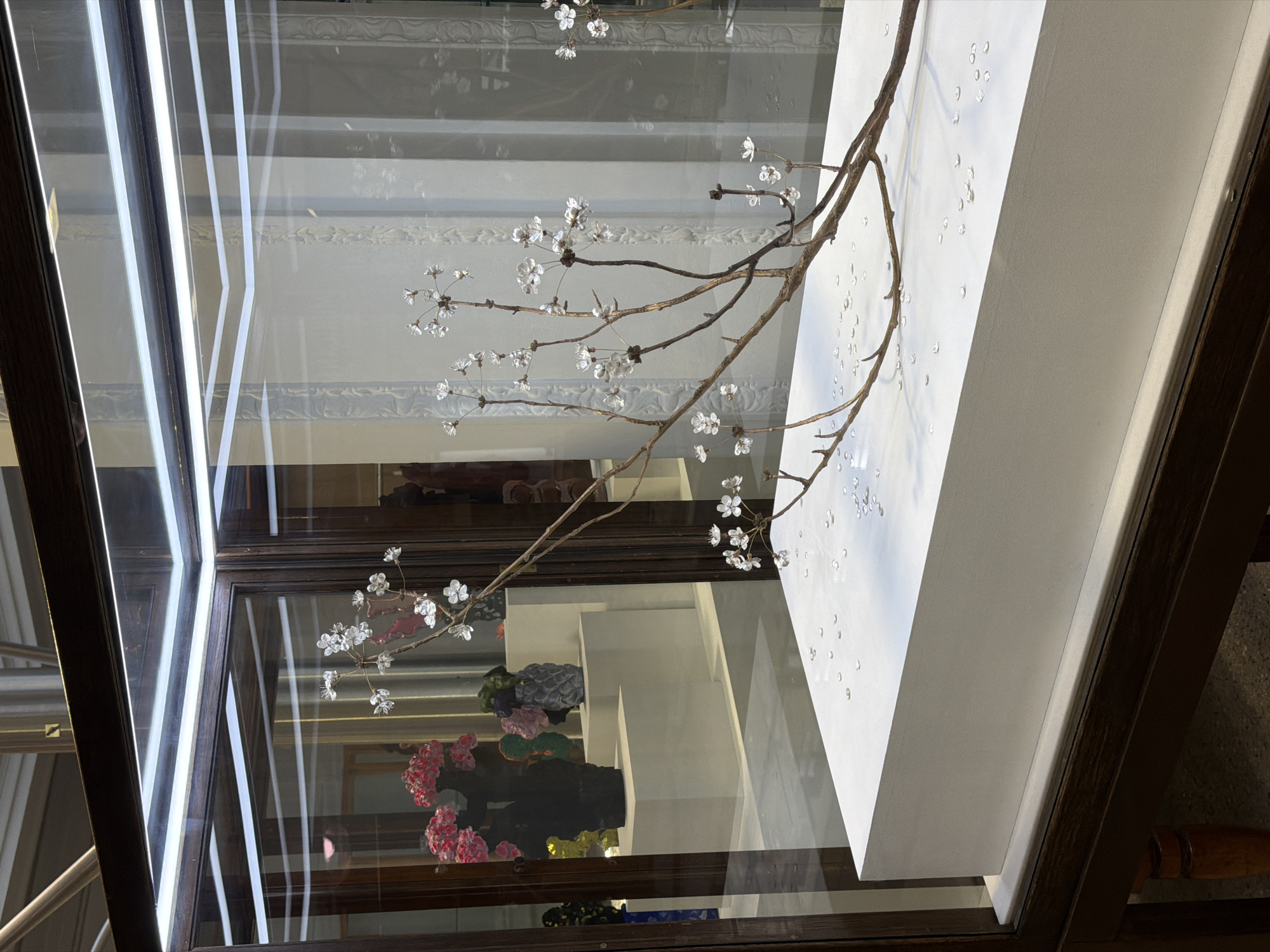 The image depicts a display case, likely located in the city of London, United Kingdom. The case is illuminated by bright, linear lights, creating stark reflections on the glass surface. Inside, a stylized branch with small, crystalline flowers is visible. The branch extends across the display, with some elements positioned against a white, angled surface. Below, partially visible through the case, are other decorative items, including floral arrangements in vases or on stands, showcasing a range of colors. The display case has a dark, possibly wooden frame. The background is a blur of interior architectural details, adding depth to the scene.