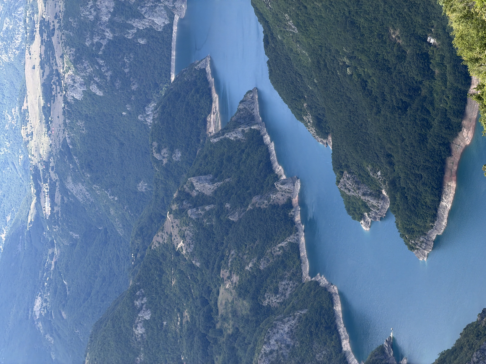 The image is an aerial view of a deep, winding river canyon in Dubljevići, Montenegro. The canyon walls are steep and covered in dense green forestation. The river itself is a vibrant blue, reflecting the sky. Rocky outcrops and bare patches of earth are visible along the riverbanks and on the upper slopes of the canyon. In the distance, the landscape opens up to rolling hills with sparse vegetation and scattered human settlements, suggesting a more arid environment beyond the immediate canyon. The sky is hazy, giving the scene a soft, atmospheric quality. There are no people or explicit objects of human construction clearly visible within the canyon itself, apart from what appear to be a few very small, distant structures on the upper plateau.