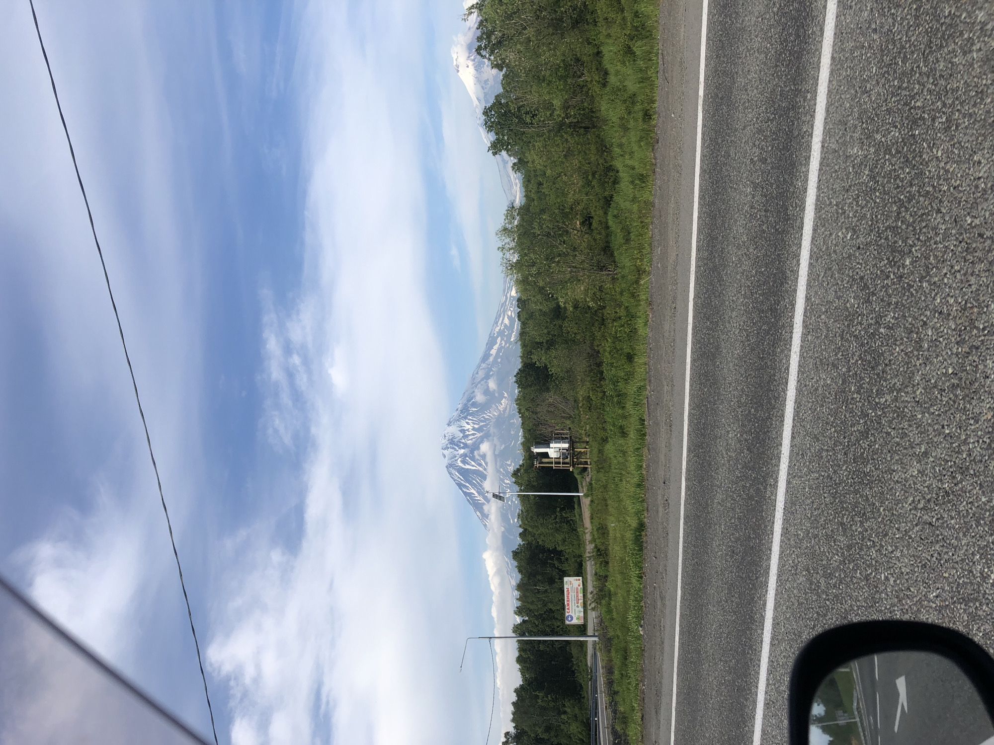 In the city of Svetly, Russia, a scenic view unfolds, dominated by majestic snow-capped volcanoes in the distance. The foreground features a paved road with white lane markings, partially visible in the lower portion of the frame. A car's side mirror, reflecting a portion of the road and greenery, is positioned in the bottom left corner. To the right of the road, a grassy embankment leads to a line of lush green trees. Beyond the trees, a large volcano, its peak dusted with snow and clouds clinging to its slopes, stands prominently against a partly cloudy blue sky. Another smaller, snow-capped volcano is visible to the right of the main one. In the middle ground, a lamppost and a solar-powered weather monitoring station are situated near the trees. A billboard with Russian text and images of food items is also visible on the left side of the scene. The overall impression is one of natural beauty and a tranquil, possibly rural or suburban, environment.