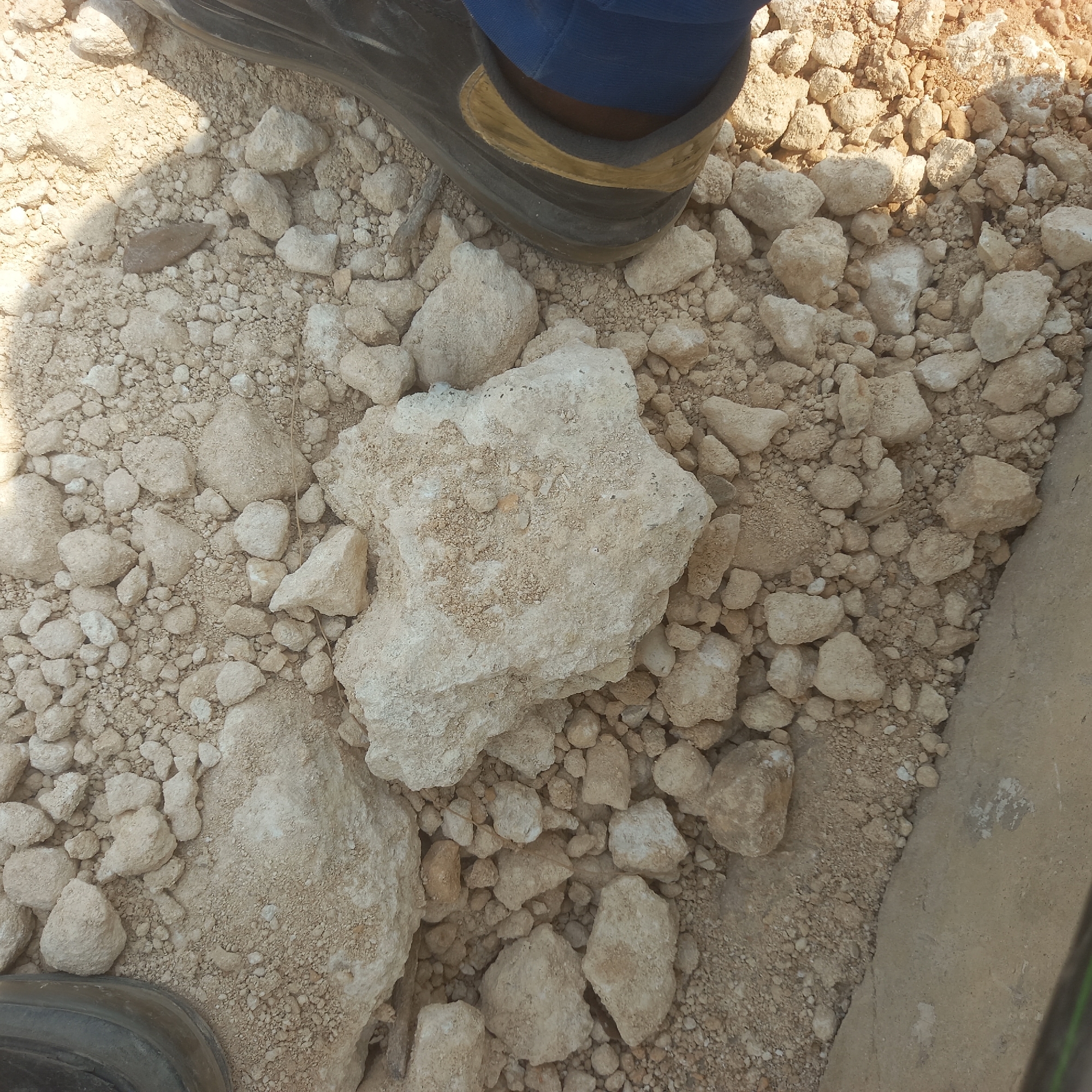 The image shows a close-up view of light-colored, irregular stones and gravel on a dirt surface. Two dark-colored boots are visible, one on the left side of the frame and another on the right, partially covering the ground. The boot on the right appears to be a work boot, with blue fabric visible around the ankle. The ground is covered in a mixture of sand and larger stones, with some smaller pebbles interspersed. The lighting suggests an outdoor environment, possibly during the daytime, with shadows cast by the stones and boots. The location is described as being in Bububu, Tanzania. There are no visible people other than the feet in the boots. The scene is static, with no discernible actions or interactions taking place.