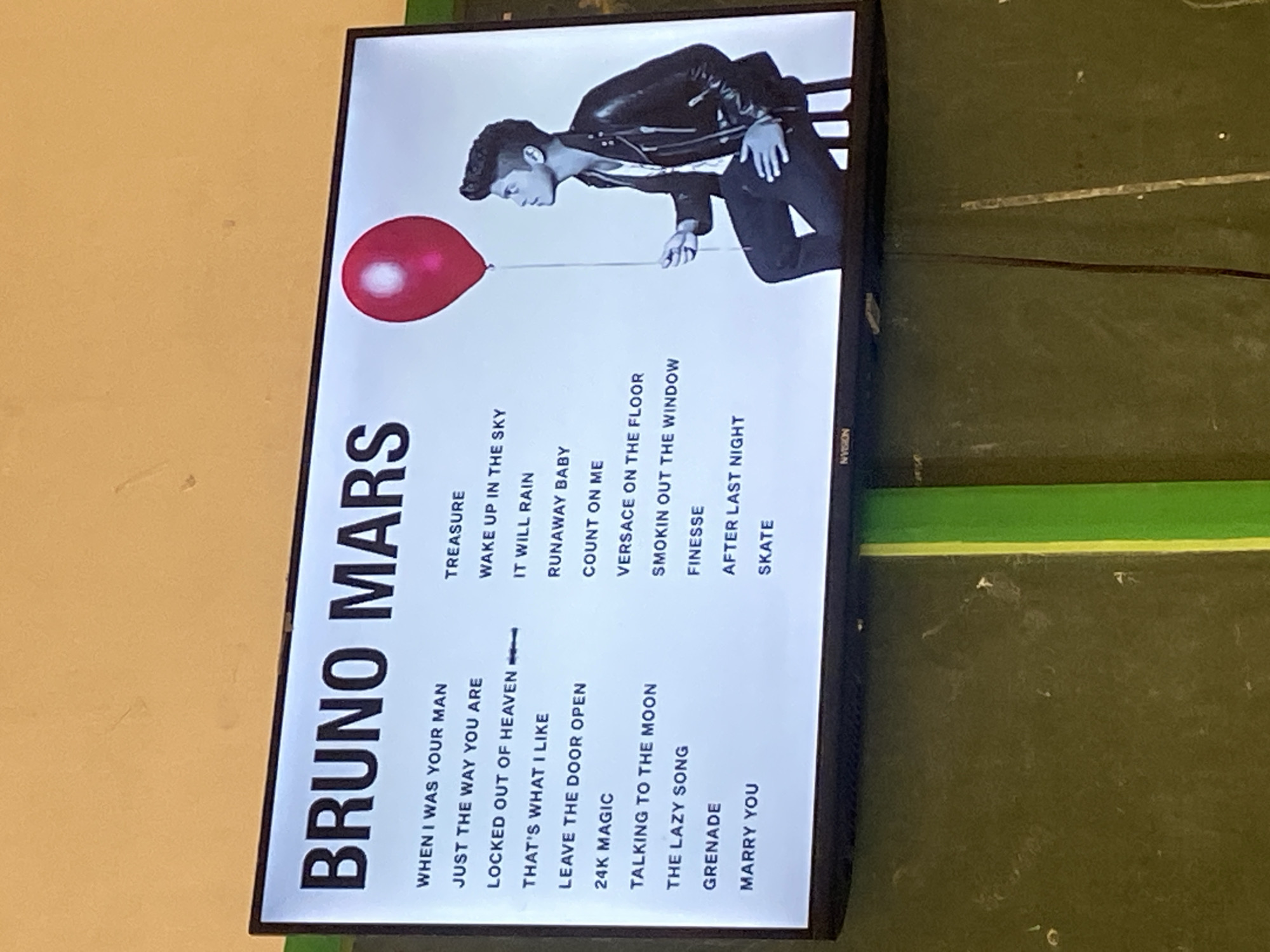 The image displays a screen showing a list of Bruno Mars songs. The screen is positioned above a dark green wall with a vertical yellow and green stripe on the left side. The screen itself is black with a white border and features the text "BRUNO MARS" in large, bold, white capital letters at the top. Below the title, two columns of song titles are listed. On the left, the songs are: "WHEN I WAS YOUR MAN", "JUST THE WAY YOU ARE", "LOCKED OUT OF HEAVEN", "THAT'S WHAT I LIKE", "LEAVE THE DOOR OPEN", "24K MAGIC", "TALKING TO THE MOON", "THE LAZY SONG", "GRENADE", and "MARRY YOU". On the right, the songs are: "TREASURE", "WAKE UP IN THE SKY", "IT WILL RAIN", "RUNAWAY BABY", "COUNT ON ME", "VERSACE ON THE FLOOR", "SMOKIN OUT THE WINDOW", "FINESSE", "AFTER LAST NIGHT", and "SKATE". To the right of the song lists, there is an image of a man, presumably Bruno Mars, holding a red balloon and sitting on a stool. The man is wearing a black leather jacket, a white t-shirt, and dark pants. The background of the screen is white. The screen has a visible brand name "NVISION" at the bottom center. The environment appears to be an indoor setting, possibly a school or community hall in Panabo City, Philippines, given the wall's appearance.