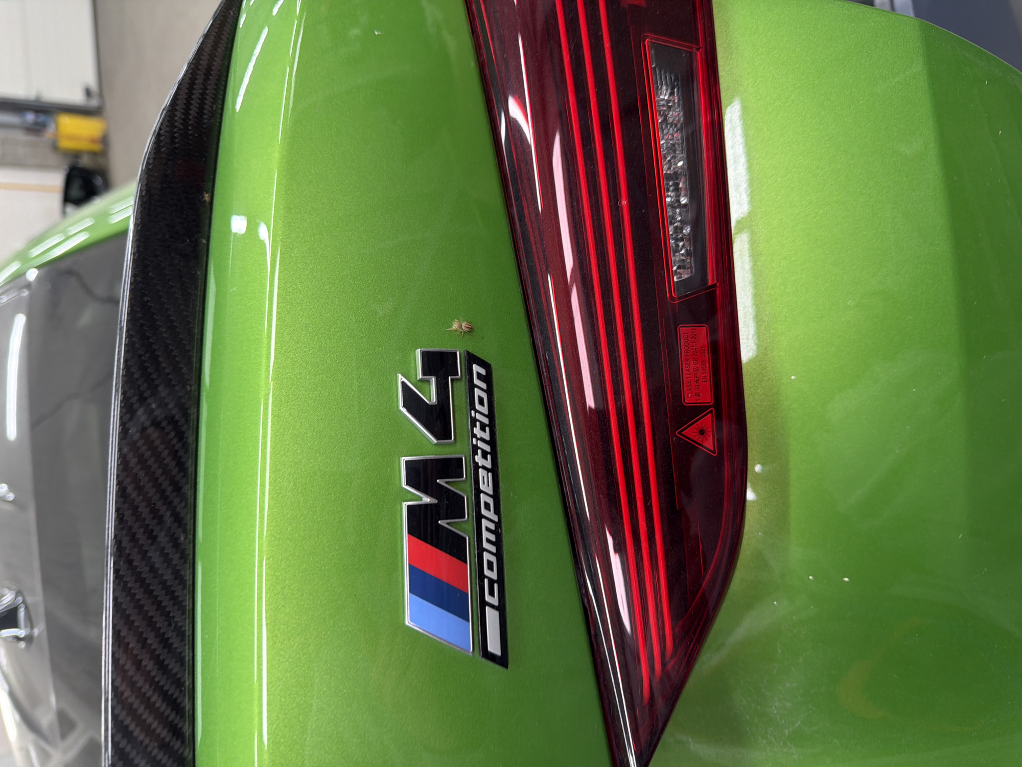 Here is a detailed description of the image:
The image shows a close-up of the rear end of a green BMW M4 Competition. The M4 Competition badge is visible, along with the edge of a carbon fiber spoiler and the car's taillight. A small insect is perched on the car's body near the badge. The scene appears to be indoors, possibly in a garage or showroom, based on the lighting and the background details. The make and model of the car are prominently displayed, highlighting the vehicle's brand and specifications.