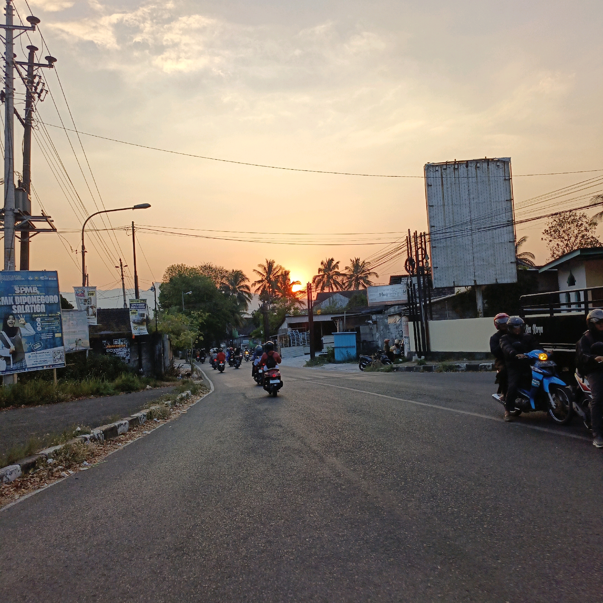 The image depicts a street scene in Salatiga, Indonesia, during sunset. The sky is illuminated with warm hues of orange and yellow, with a few clouds visible. A road runs diagonally from the bottom left to the top right of the frame, carrying traffic primarily consisting of motorcycles. Several motorcycles with riders are visible on the road, some in motion and others stopped.
On the left side of the road, there is a large billboard displaying information about "SPMK DIPONEGORO BUSINESS SCHOOL" and "SMK DIPONEGORO SALATIGA". A utility pole with multiple wires runs alongside the billboard. Further down the road on the left, there are buildings and trees. On the right side of the road, a tall, blank advertising billboard stands out, with electrical wires crisscrossing in front of it. Adjacent to this billboard are more buildings, some appearing to be shops or residences, and more utility poles and wires. Palm trees are visible silhouetted against the sunset sky, particularly in the middle distance. The overall environment suggests an urban or semi-urban setting. The road itself is asphalt, with a concrete curb and some vegetation along its edge. The time of day is clearly sunset, indicated by the low angle of the sun and the vibrant colors in the sky.