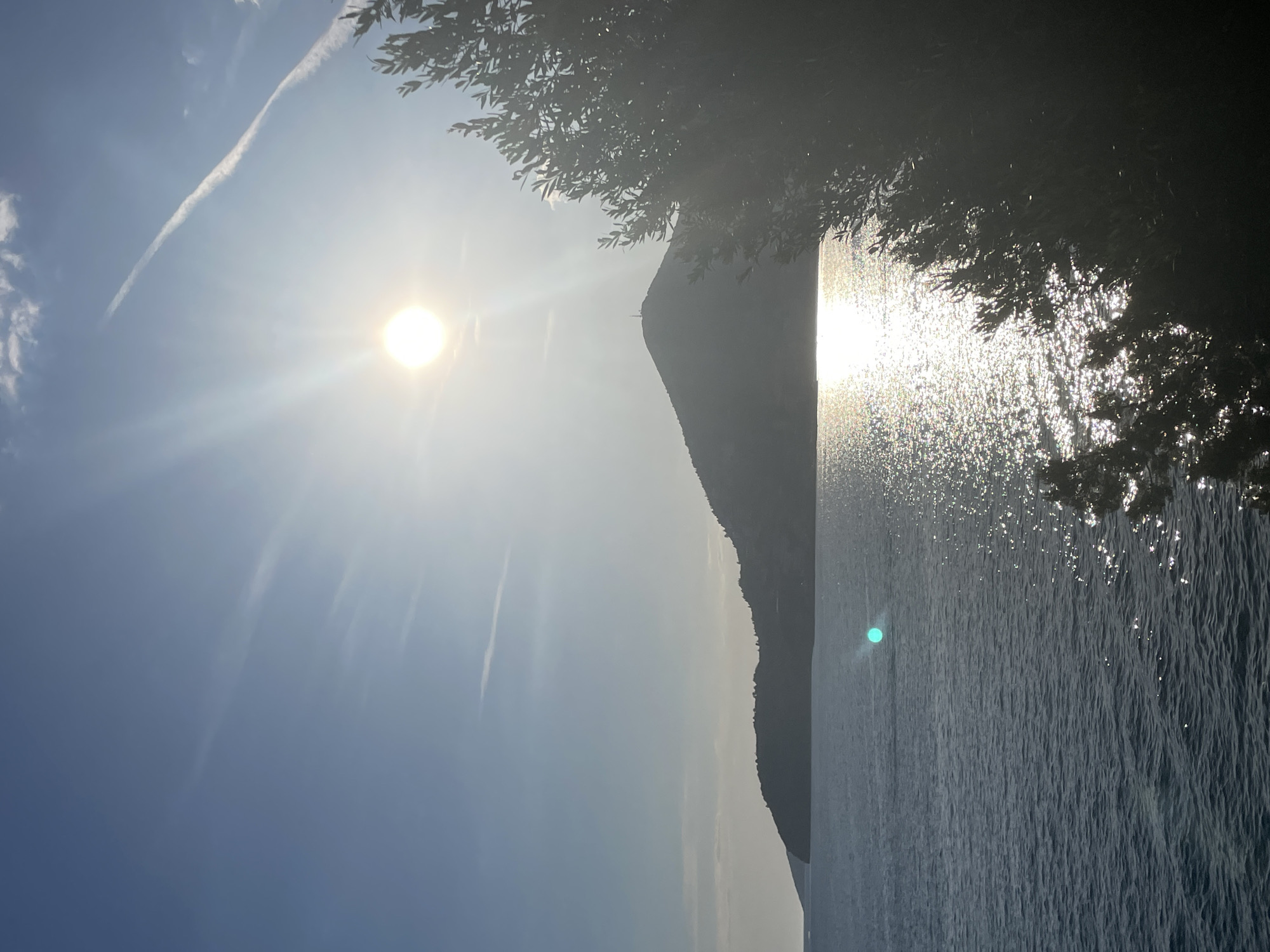The image captures a daytime scene in Boreti, Montenegro, with a focus on the interplay of sunlight and the natural landscape. The sun is prominently positioned in the upper left quadrant, its rays spreading across the sky and reflecting intensely on the water. The body of water, likely the Adriatic Sea, extends vertically on the right side of the frame, its surface shimmering with sunlight. A mountain silhouette forms a vertical line between the sky and the sea, adding depth to the composition. In the top right corner, tree foliage is visible, partially obscuring the view. A contrail cuts across the sky in the upper left.