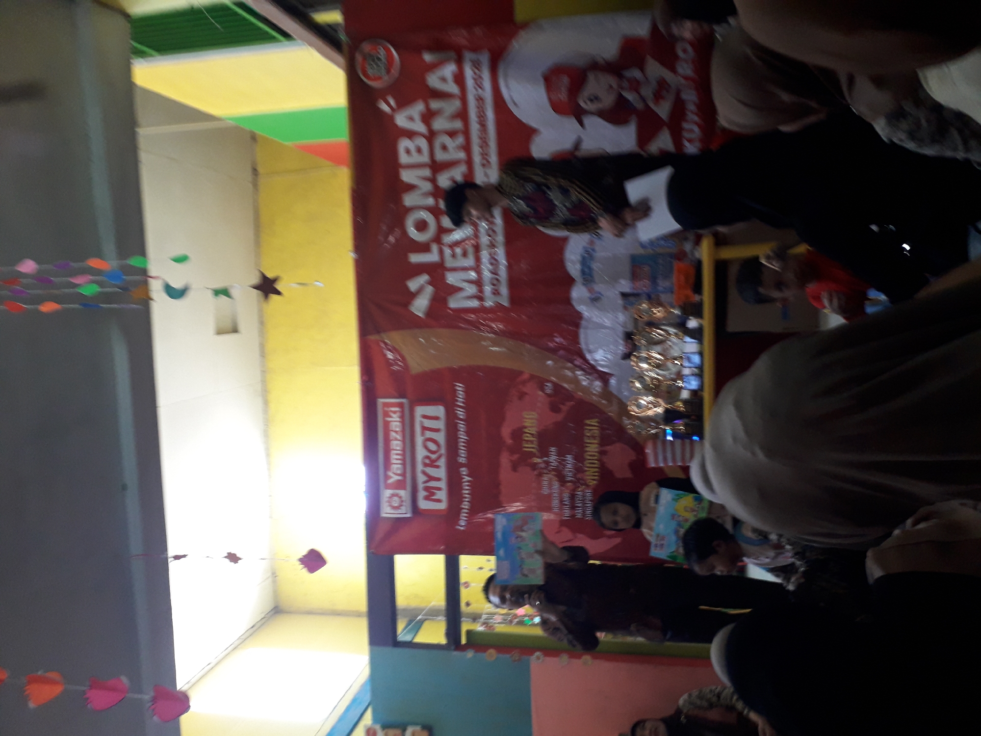 An indoor event is taking place, likely a children's coloring competition, as indicated by the banner reading "LOMBA MEWARNAI" (Coloring Competition) and the presence of young children. The scene is set with a red banner as a backdrop, featuring logos for "Yamazaki MYROTI" and "KUYROTI." A man in a dark shirt is holding a children's book and a microphone, possibly addressing the audience or the child he is interacting with. A young girl in a hijab is seated in front of him, also holding a book. Several trophies are displayed on a table to the right, suggesting prizes for the competition. The environment appears to be a community hall or school, with yellow and white walls visible. Decorations in the form of colorful paper cutouts shaped like hearts and stars hang from the ceiling. The attendees are seated, with many wearing headscarves. The overall atmosphere seems to be festive and engaging for children. The event appears to be held in December 2020.